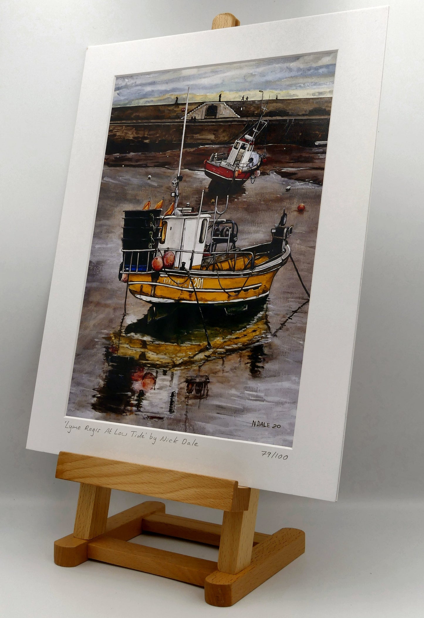 Image of Medium Art Print featuring a copy of the painting 'Lyme Regis at Low Tide' by Nick Dale. The print depicts a sunset where the fishing vessels rest on the floor of Lyme Regis harbour whilst the tourists enjoy the view from the Cobb. The print is situated on a small easel.