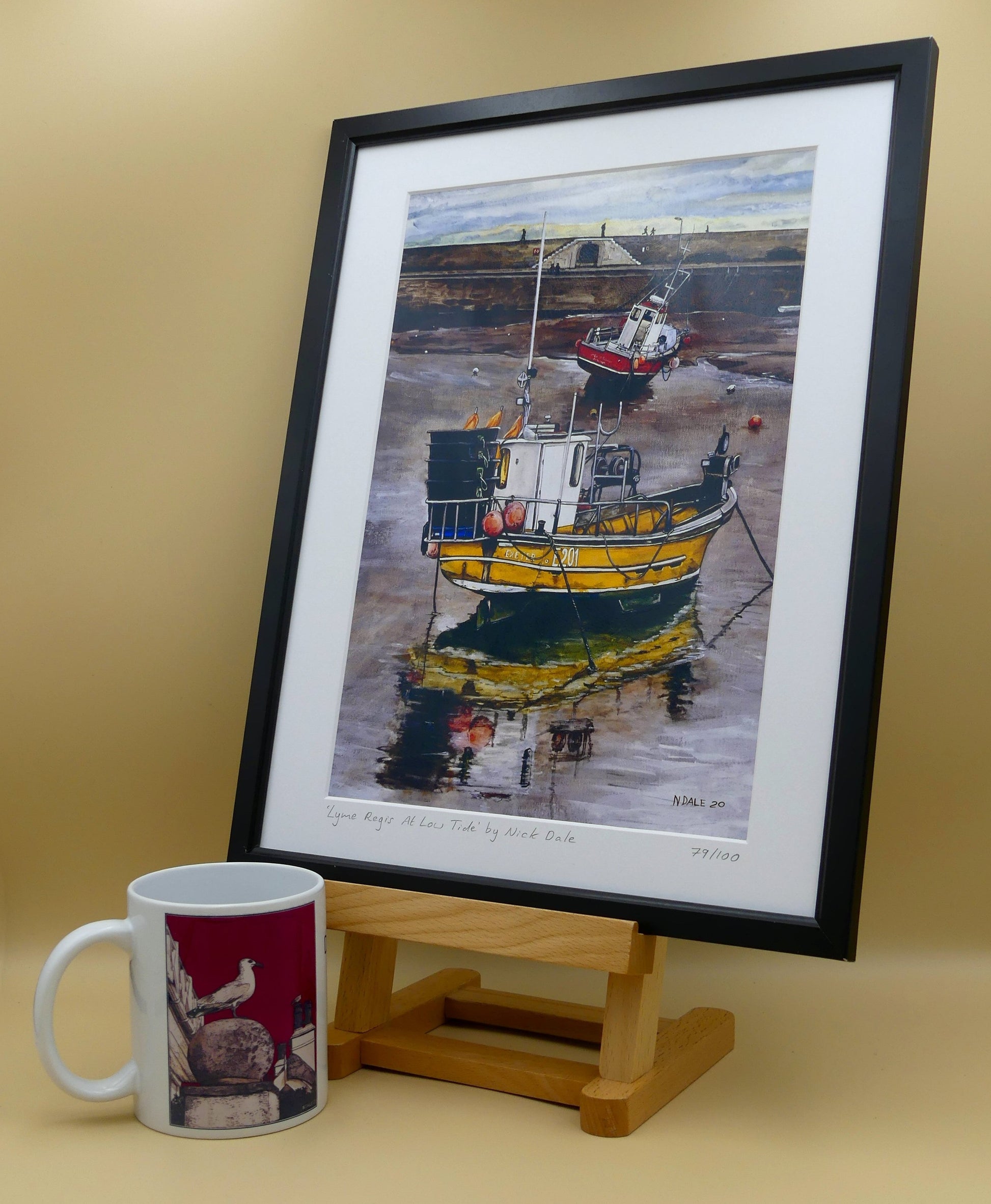 Image of Medium Art Print featuring a copy of the painting 'Lyme Regis at Low Tide' by Nick Dale. The print depicts a sunset where the fishing vessels rest on the floor of Lyme Regis harbour whilst the tourists enjoy the view from the Cobb. The print is situated in a black frame next to a mug.