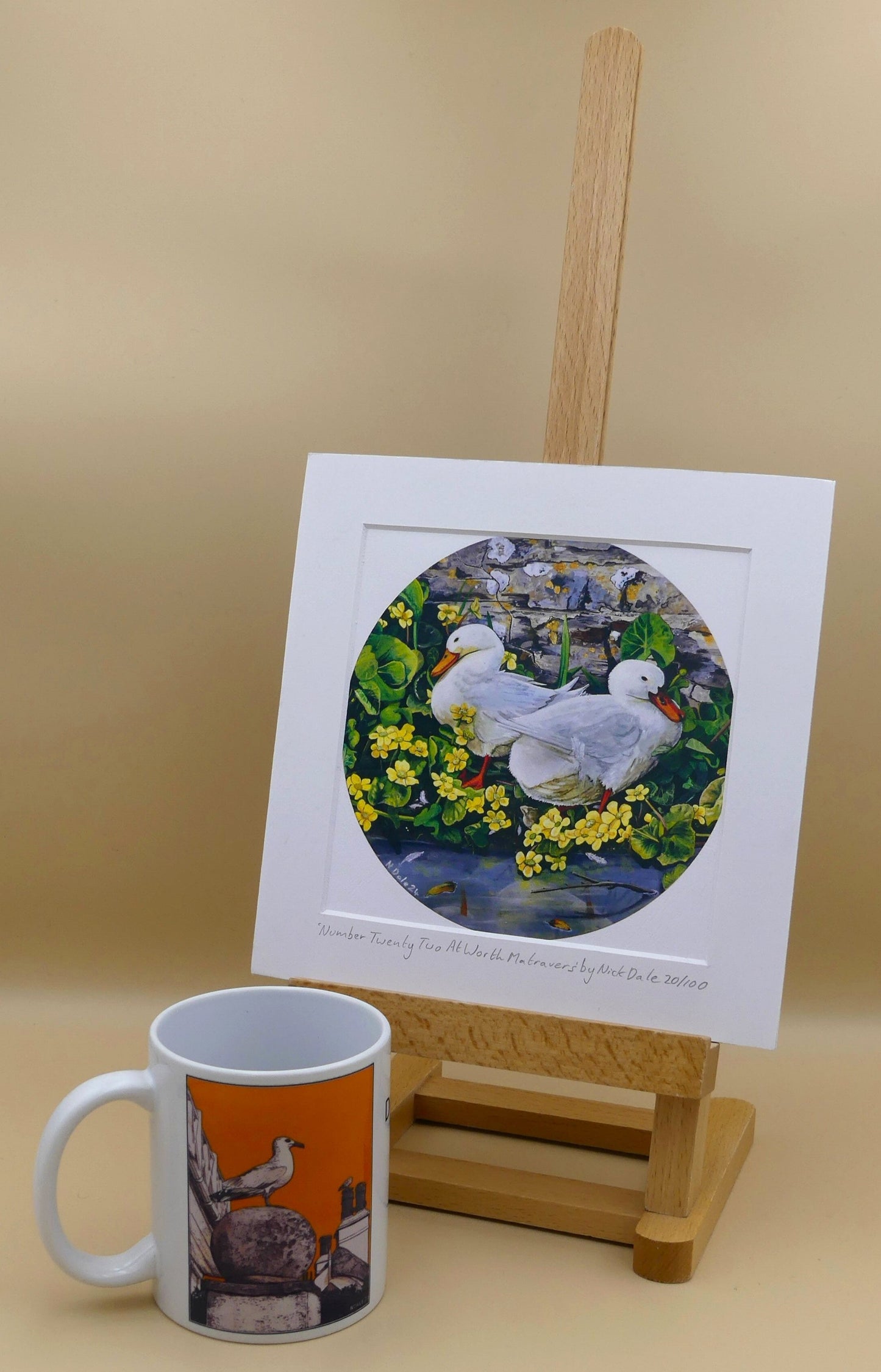 Image of Square Medium Art Print featuring a copy of the painting 'Number Twenty Two At Worth Matravers' by Nick Dale. The print depicts 2 domestic ducks on the edge of the duck pond at Worth Matravers in Dorset. 'Two Little Ducks' is often used as the call in bingo for the number 22 - hence the title of the painting. The print is situated on a small easel next to a mug.