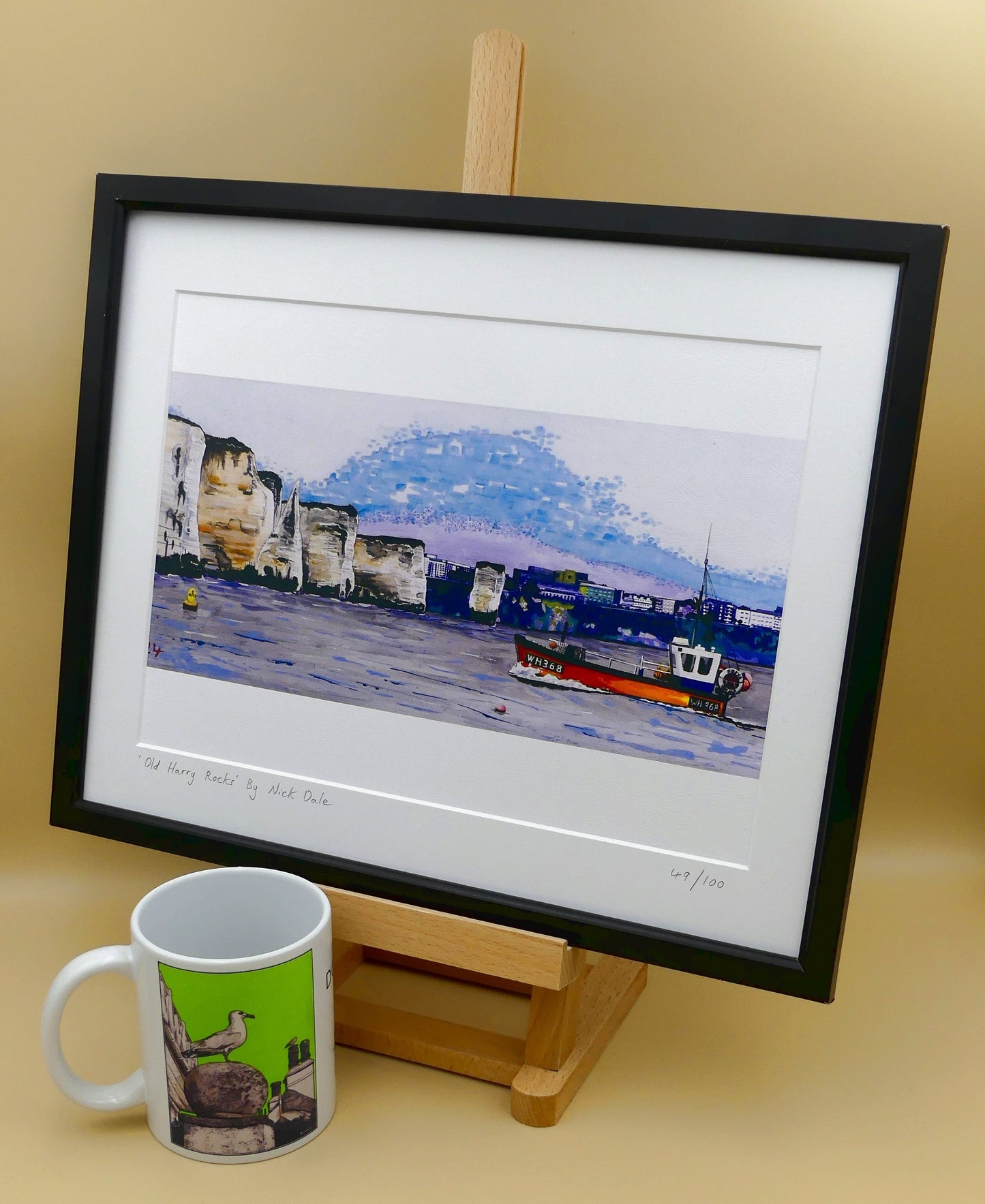 Image of a Medium Art Print (A4 size) featuring a copy of a detail of the painting 'Old Harry Rocks' by Nick Dale. The print depicts the chalk formations located at Handfast Point, south of Studland Bay; as viewed from Peveril Point, Swanage. Also depicted is a small fishing boat returning to Swanage and the Bournemouth skyline beyond the cliffs. The print is situated on a small easel next to a mug.