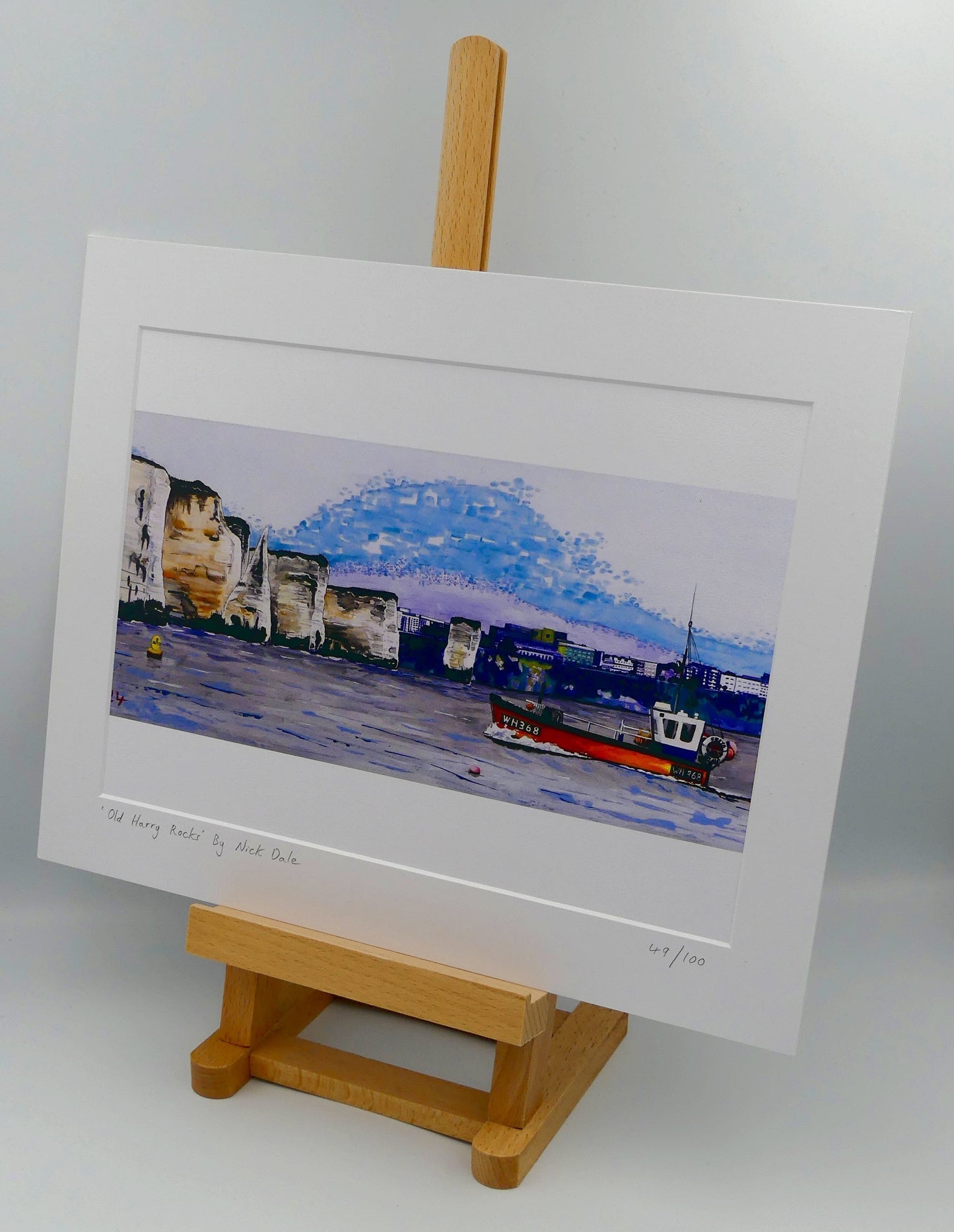 Image of a Medium Art Print (A4 size) featuring a copy of a detail of the painting 'Old Harry Rocks' by Nick Dale. The print depicts the chalk formations located at Handfast Point, south of Studland Bay; as viewed from Peveril Point, Swanage. Also depicted is a small fishing boat returning to Swanage and the Bournemouth skyline beyond the cliffs. The print is situated on a small easel.
