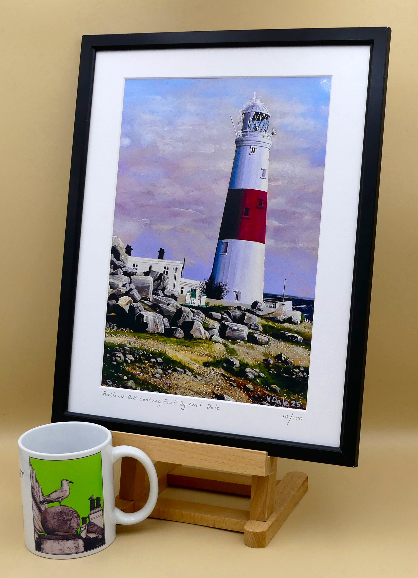 Image of a Medium Art Print featuring a copy of the painting 'Portland Bill Looking East' by Nick Dale. The print depicts a view of Portland Bill Lighthouse as seen from the quarry area to the west, looking east. The print is situated on a small easel in a black frame next to a mug.