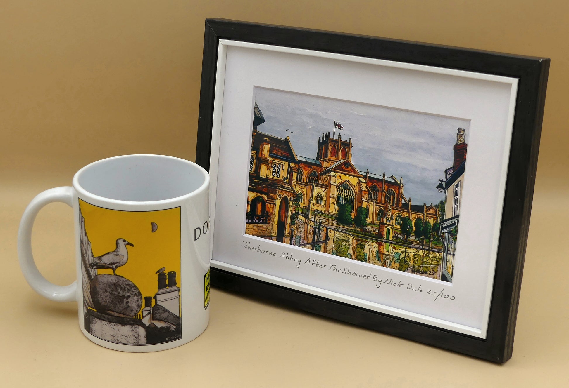 Image of Small Art Print featuring a copy of the painting 'Sherborne Abbey After The Shower' by Nick Dale. The print depicts a view of Sherborne Abbey (also known as the Abbey Church of St. Mary the Virgin), in Sherborne, Dorset. On the left hand side can be seen St Johns' Almshouse.  The print is situated in a black frame next to a mug.