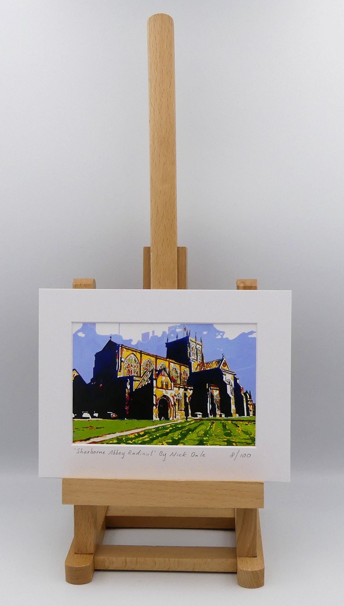 Image of a Small Art Print featuring a copy of the painting 'Sherborne Abbey Radiant' by Nick Dale. The print depicts Sherborne Abbey in bright primary colours with strong shadows and a brightly lit south-facing aspect. The print is situated on a small easel.