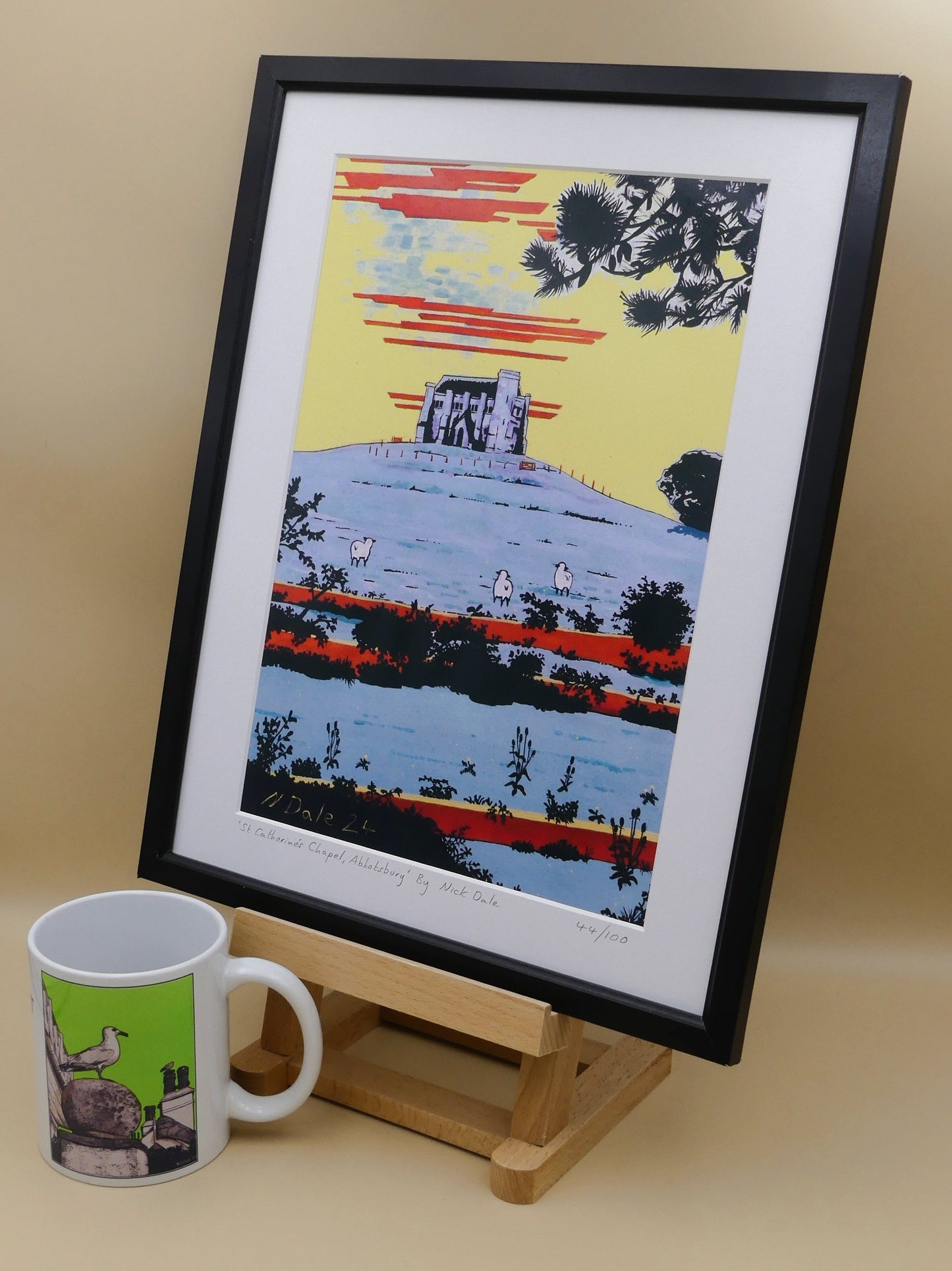 Image of a Medium Art Print (A4 size) featuring a copy of the painting 'St Catherine's Chapel, Abbotsbury' by Nick Dale. The print depicts the Medieval St Catherine's Chapel which is positioned overlooking the village of Abbotsbury. The print is situated on a small easel next to a mug.