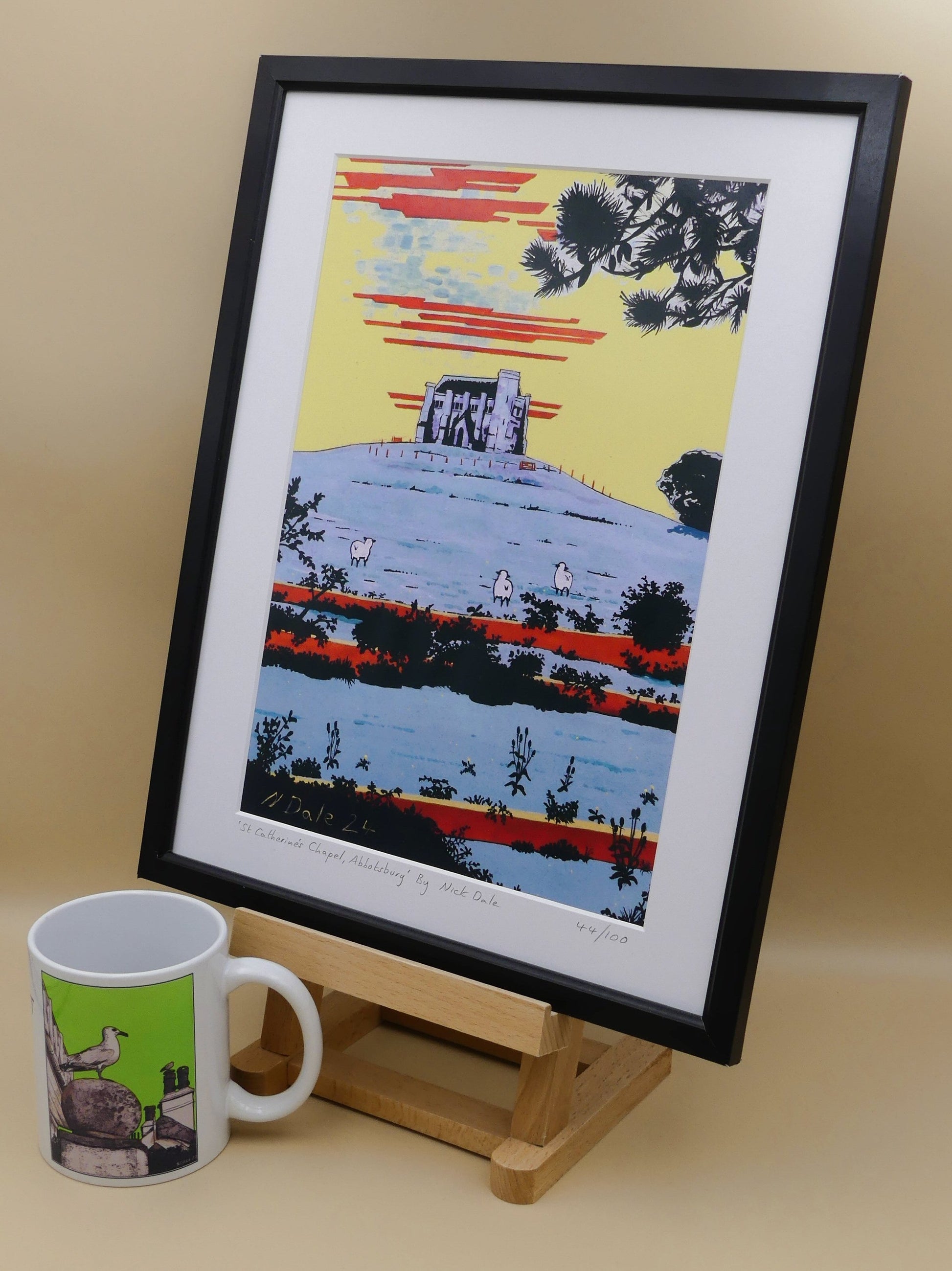 Image of a Medium Art Print (A4 size) featuring a copy of the painting 'St Catherine's Chapel, Abbotsbury' by Nick Dale. The print depicts the Medieval St Catherine's Chapel which is positioned overlooking the village of Abbotsbury. The print is situated on a small easel next to a mug.