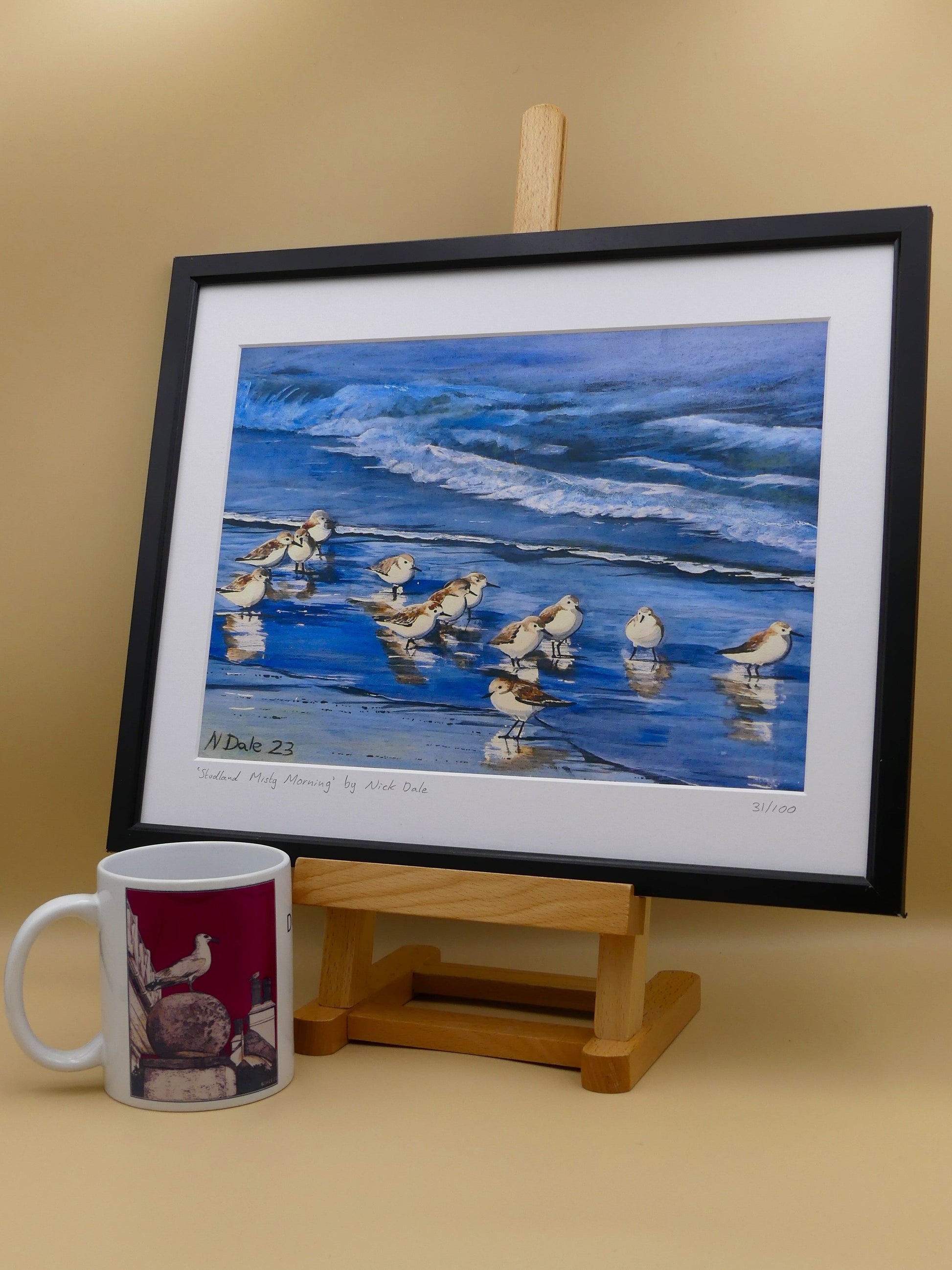 Image of a Medium Art Print featuring a copy of the painting 'Studland Misty Morning' by Nick Dale. The print depicts sanderlings on Studland Beach (Dorset) on a crisp and misty morning. The print is situated in a black frame next to a mug.