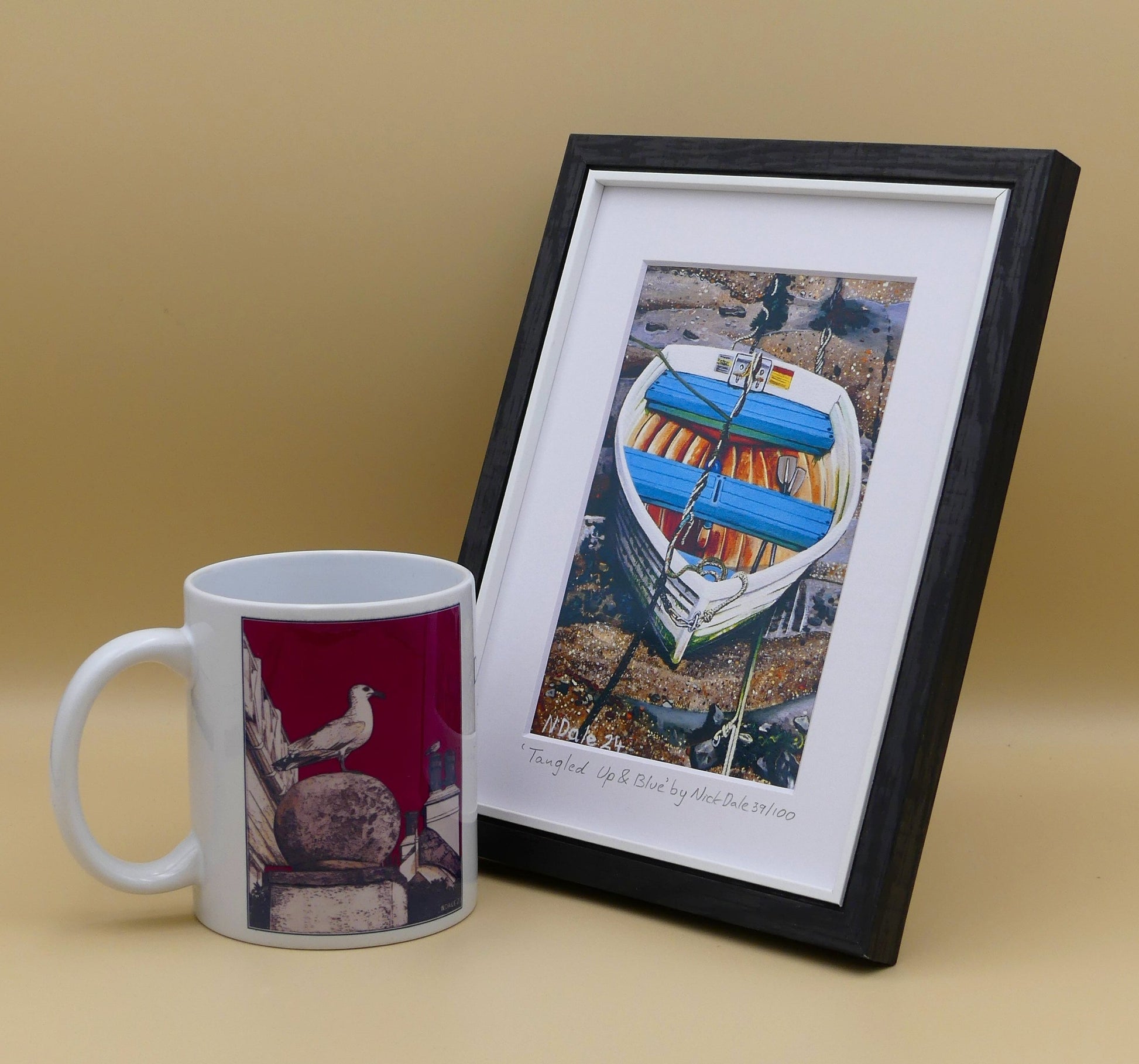 Image of a Small Art Print featuring a copy of the painting 'Tangled Up & Blue' by Nick Dale. The print depicts a small rowing boat sitting on the floor of Lynmouth Harbour at Lynmouth in Devon. The print is situated in a black frame next to a mug.