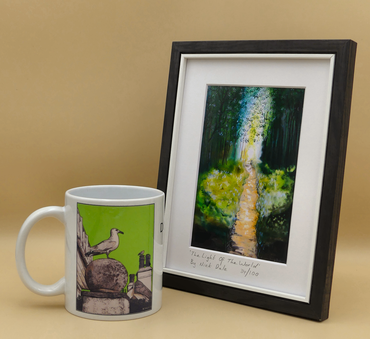 Image of a Small Art Print featuring a copy of the painting 'The Light of the World' by Nick Dale. The print depicts a woodland path at Westonbirt Arboretum (near the town of Tetbury in Gloucestershire) illuminated by the sunlight streaming through the trees. The print is situated in a black frame next to a mug.