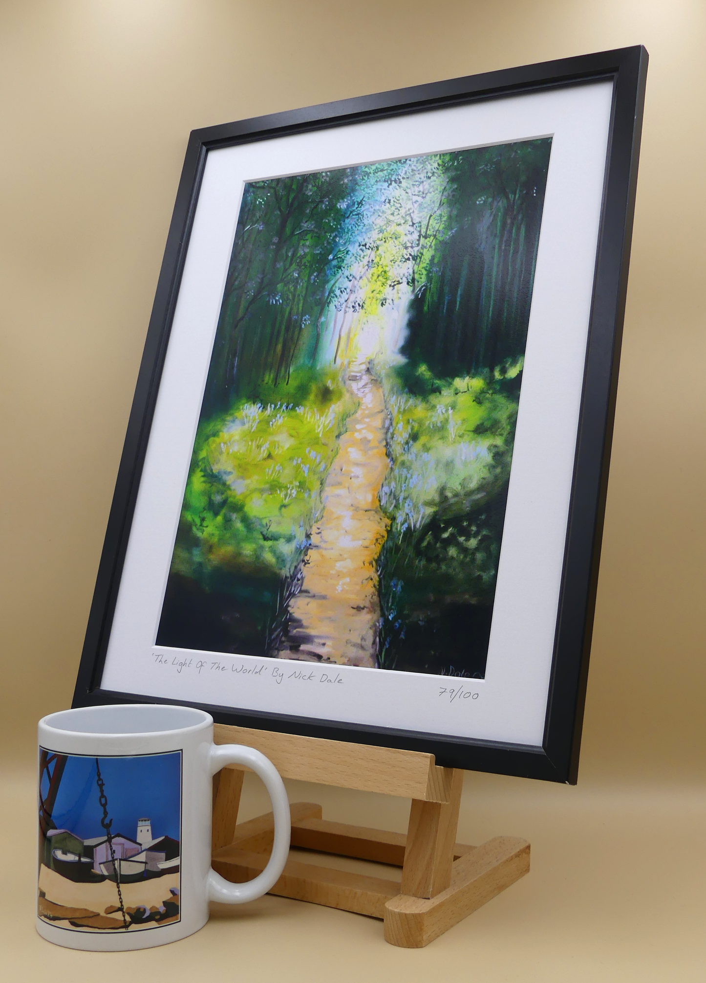 Image of a Medium Art Print featuring a copy of the painting 'The Light of the World' by Nick Dale. The print depicts a woodland path at Westonbirt Arboretum (near the town of Tetbury in Gloucestershire) illuminated by the sunlight streaming through the trees. The print is situated on a small easel in a black frame next to a mug.