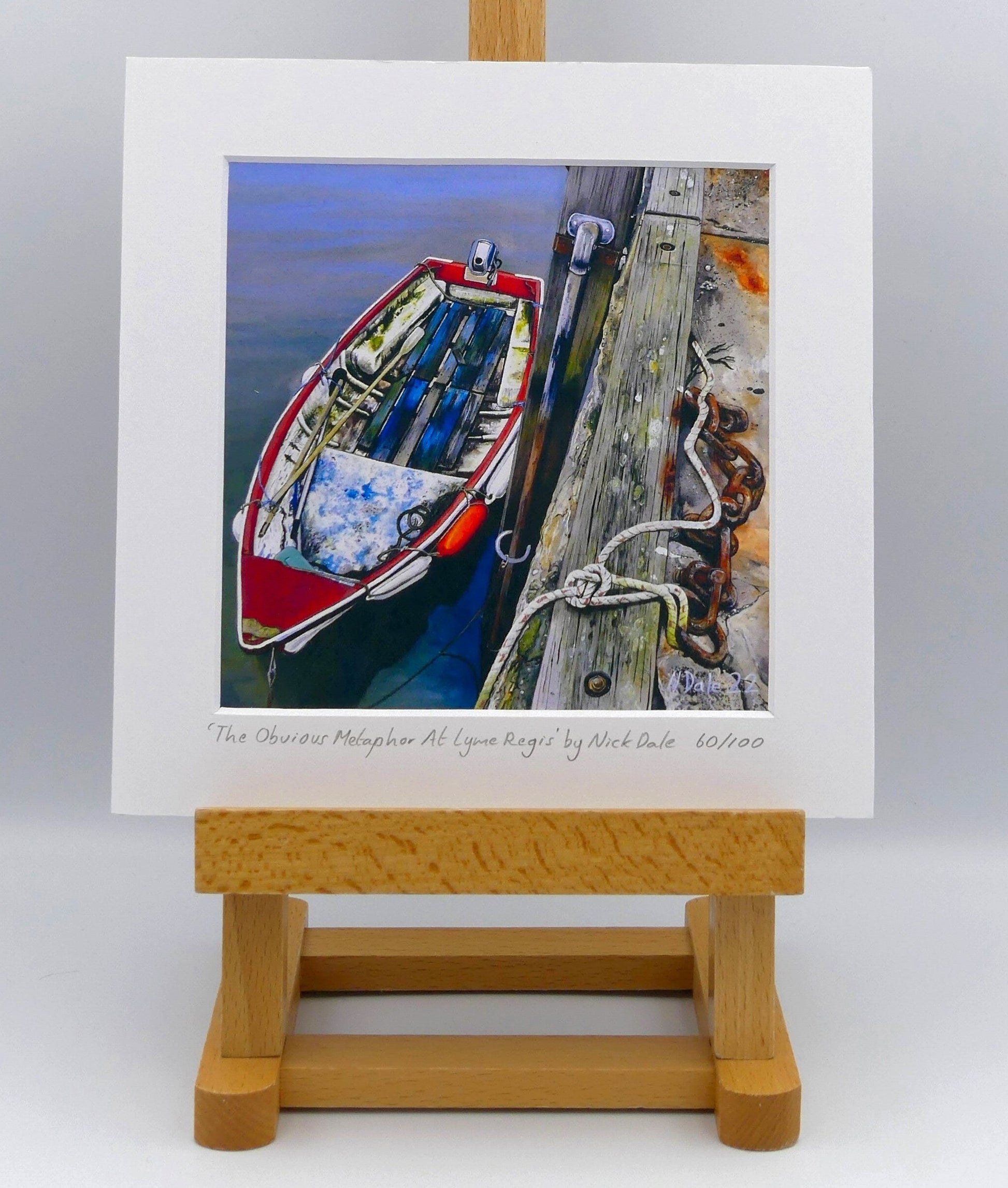 Image of Square Medium Art Print featuring a copy of the painting 'The Obvious Metaphor Of Lyme Regis' by Nick Dale. The print depicts a somewhat unkempt rowing boat aside the Cobb at Lyme Regis. The print is situated on a small easel.
