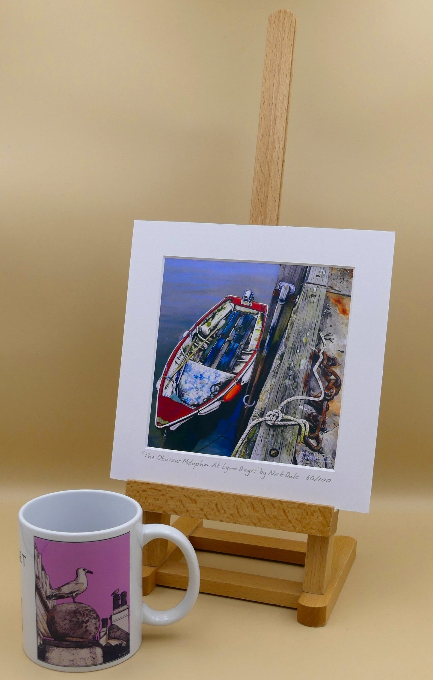 Image of Square Medium Art Print featuring a copy of the painting 'The Obvious Metaphor Of Lyme Regis' by Nick Dale. The print depicts a somewhat unkempt rowing boat aside the Cobb at Lyme Regis. As observed from Ringstead Beach. The print is situated on a small easel next to a mug.