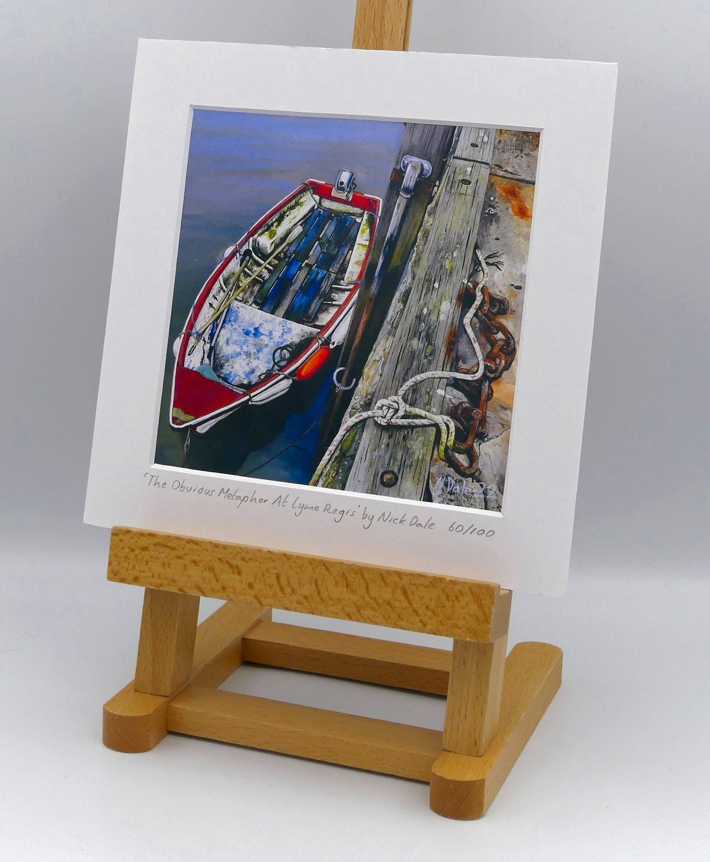Image of Square Medium Art Print featuring a copy of the painting 'The Obvious Metaphor Of Lyme Regis' by Nick Dale. The print depicts a somewhat unkempt rowing boat aside the Cobb at Lyme Regis. The print is situated on a small easel.