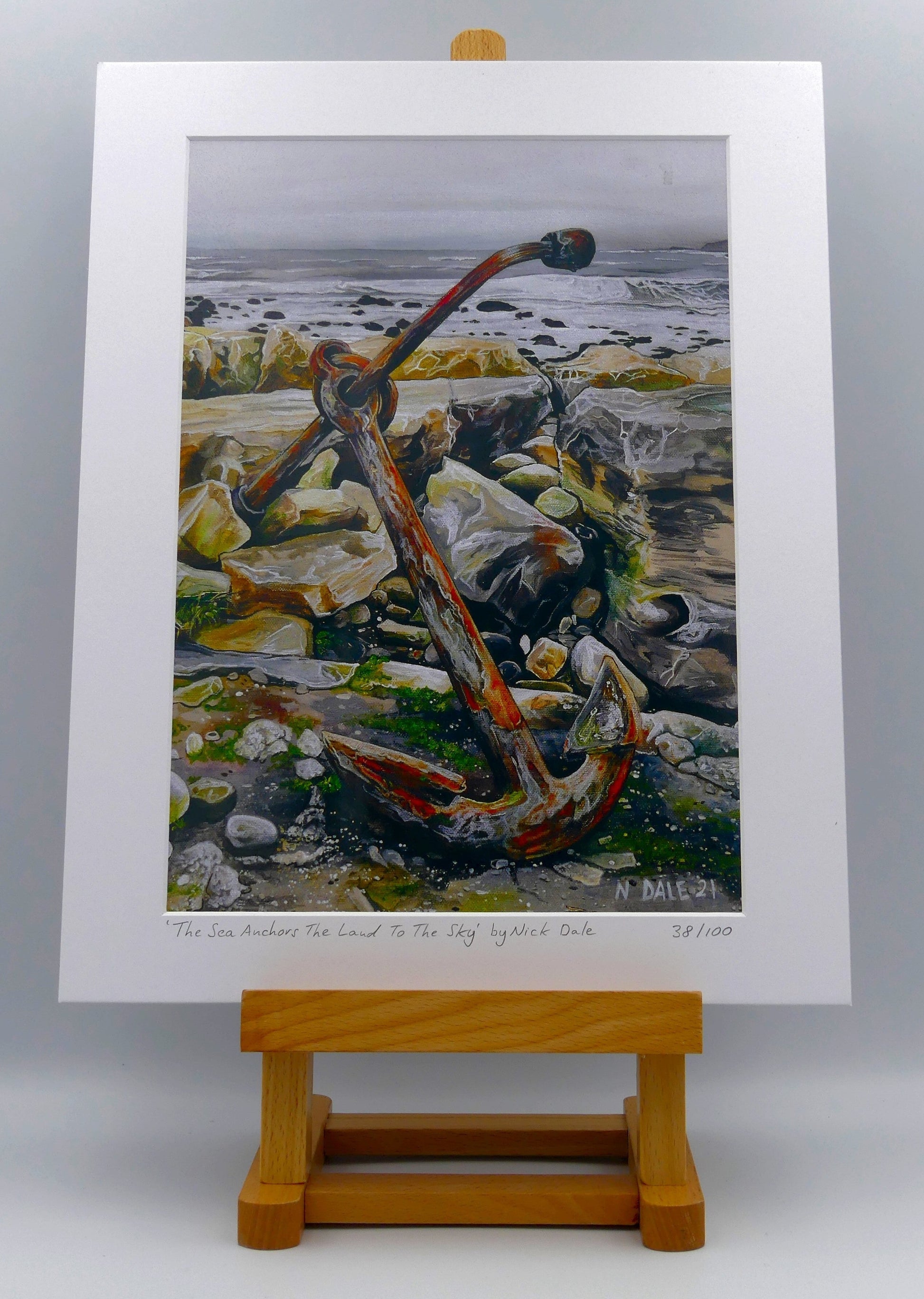 Image of a Medium Art Print featuring a copy of the painting 'The Sea Anchors The Land To The Sky' by Nick Dale. The print depicts an old anchor lying at rest under a leaden sky upon the foreshore at Chapman's Pool, near Worth Matravers, Dorset. The print is situated on a small easel.