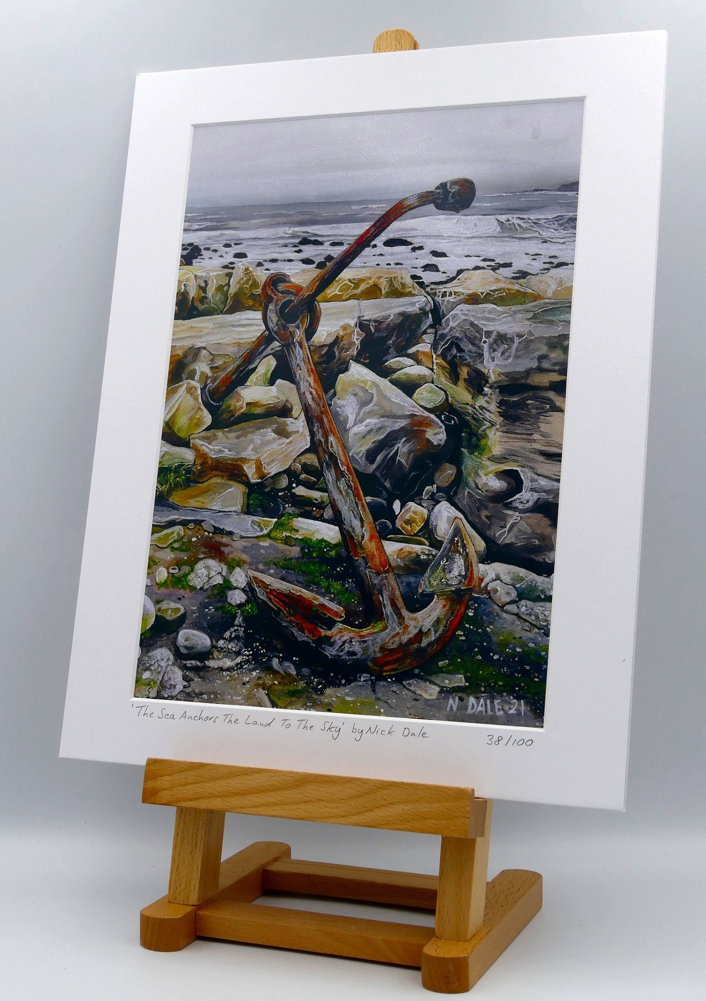Image of a Medium Art Print featuring a copy of the painting 'The Sea Anchors The Land To The Sky' by Nick Dale. The print depicts an old anchor lying at rest under a leaden sky upon the foreshore at Chapman's Pool, near Worth Matravers, Dorset. The print is situated on a small easel.