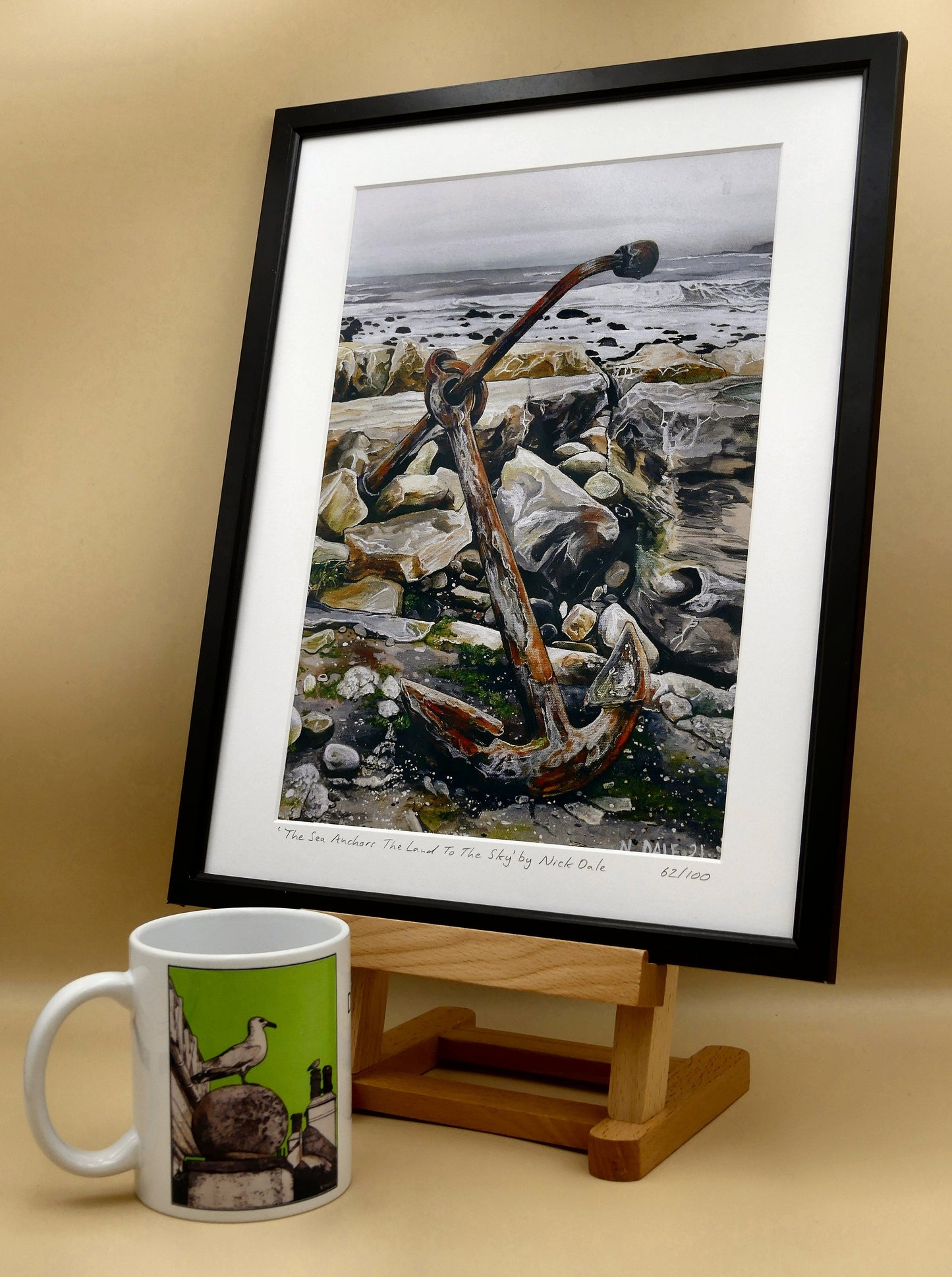 Image of a Medium Art Print featuring a copy of the painting 'The Sea Anchors The Land To The Sky' by Nick Dale. The print depicts an old anchor lying at rest under a leaden sky upon the foreshore at Chapman's Pool, near Worth Matravers, Dorset. The print is situated in a black frame next to a mug.