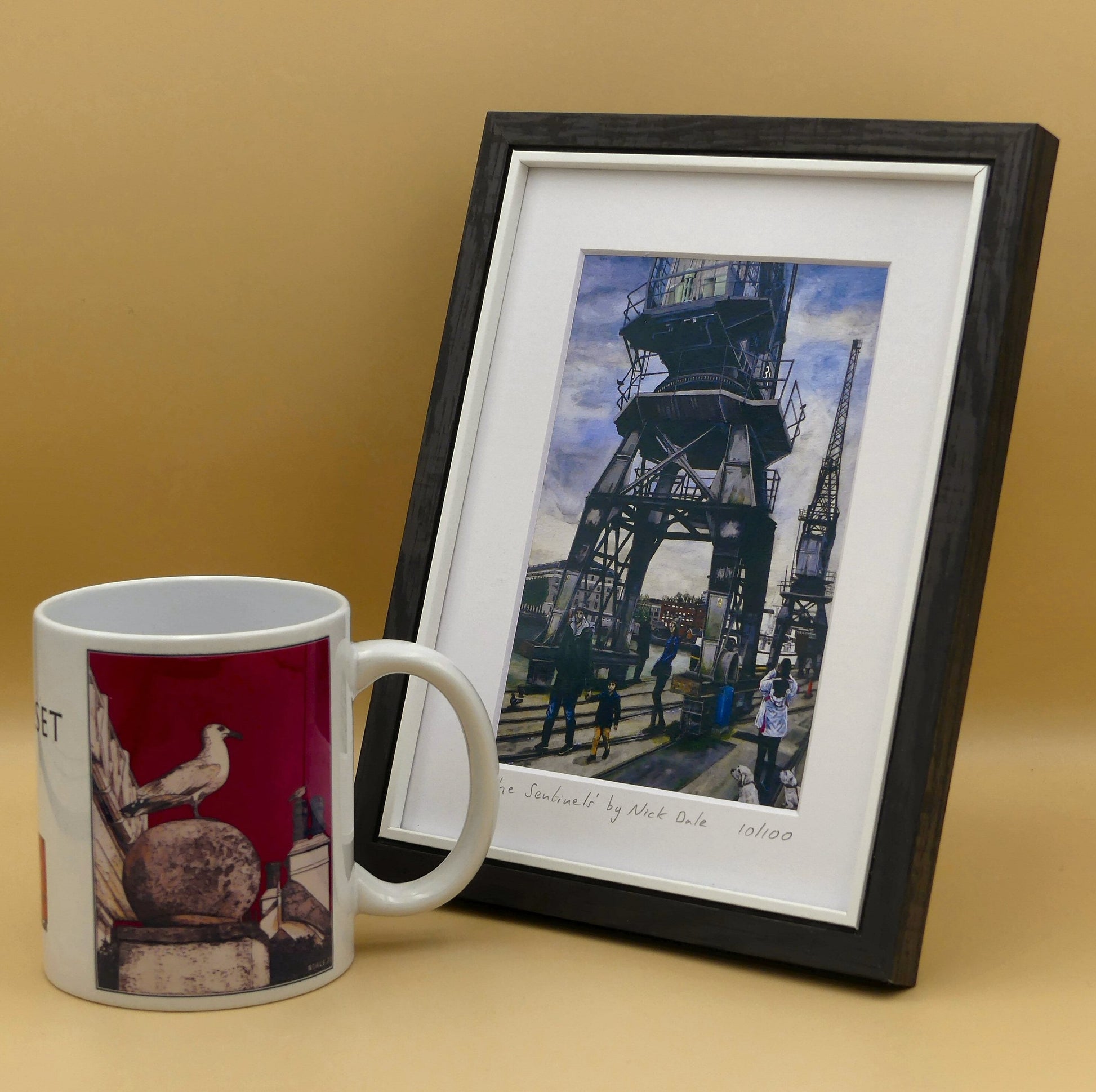 Image of a Small Art Print featuring a copy of the painting 'The Sentinels' by Nick Dale. The print depicts the iconic tower cranes of Bristol Docks and the classic ship MV Balmoral beyond them. The print is situated in a black frame next to a mug.