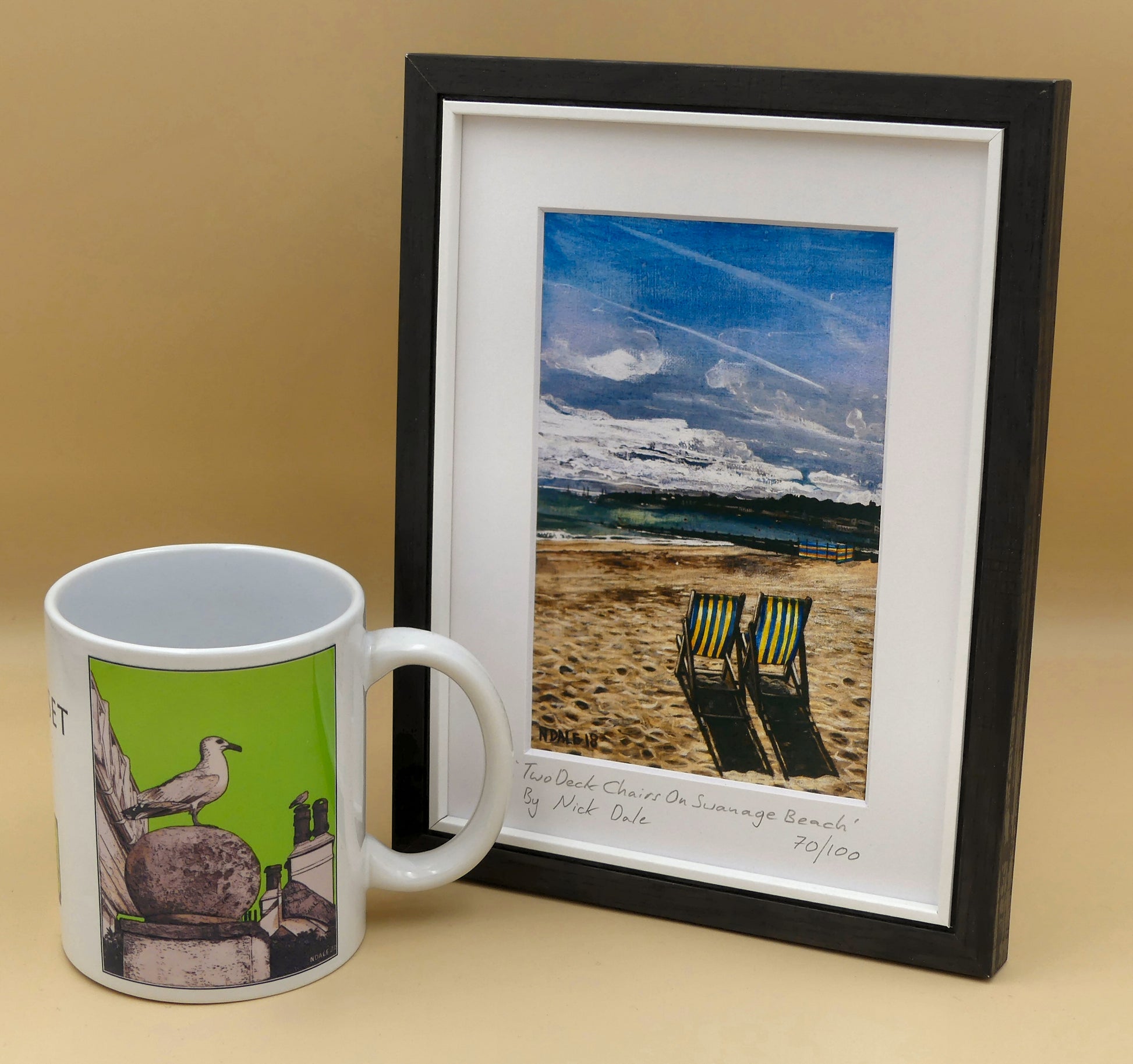 Image of a Small Art Print featuring a copy of the painting 'Two Deck Chairs On Swanage Beach' by Nick Dale. The print depicts two deck chairs overlooking the calm water from Swanage Beach (Dorset) towards Peveril Point. The print is situated in a black frame next to a mug.