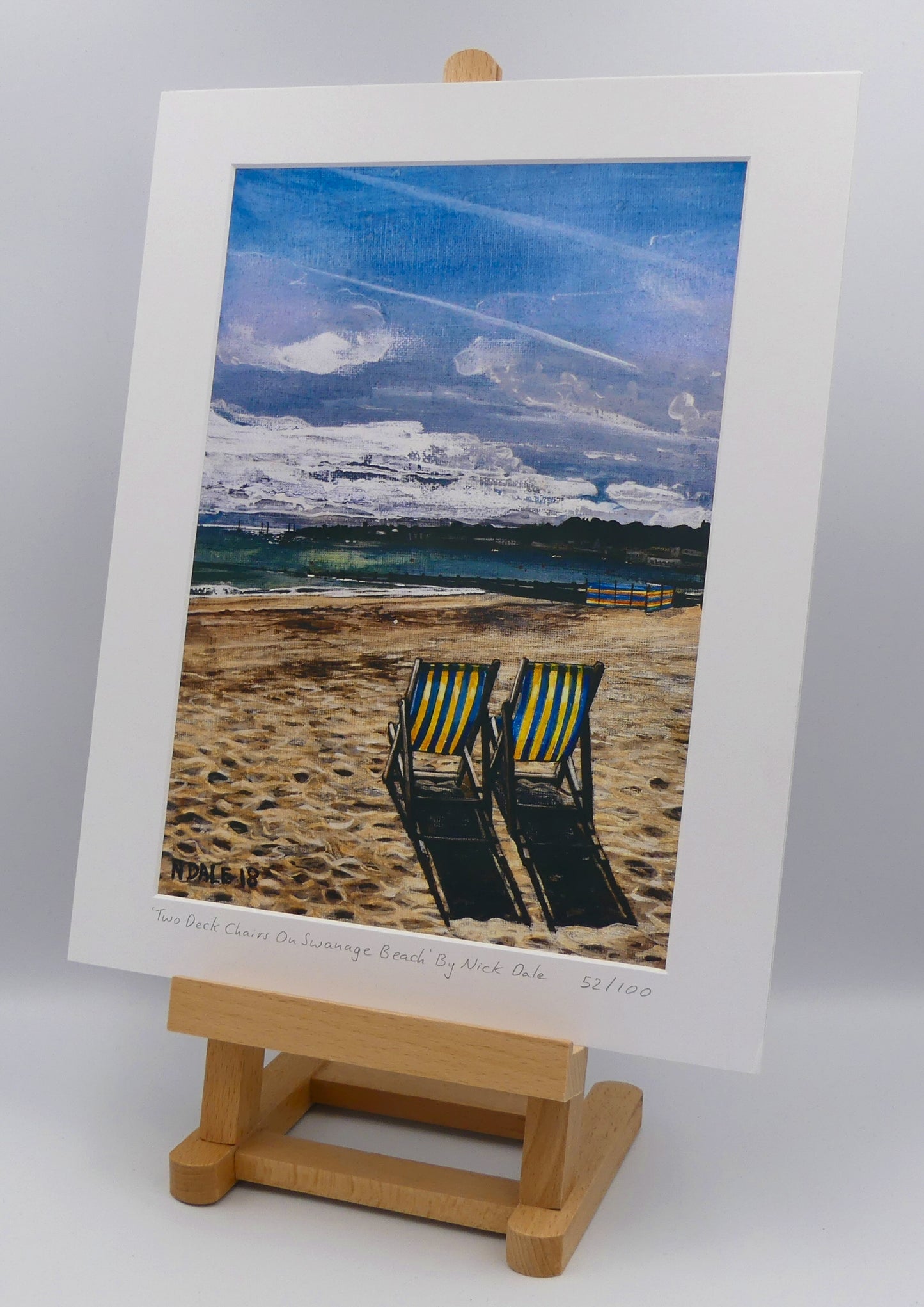 Image of a Medium Art Print featuring a copy of the painting 'Two Deck Chairs On Swanage Beach' by Nick Dale. The print depicts two deck chairs overlooking the calm water from Swanage Beach (Dorset) towards Peveril Point. The print is situated on a small easel.