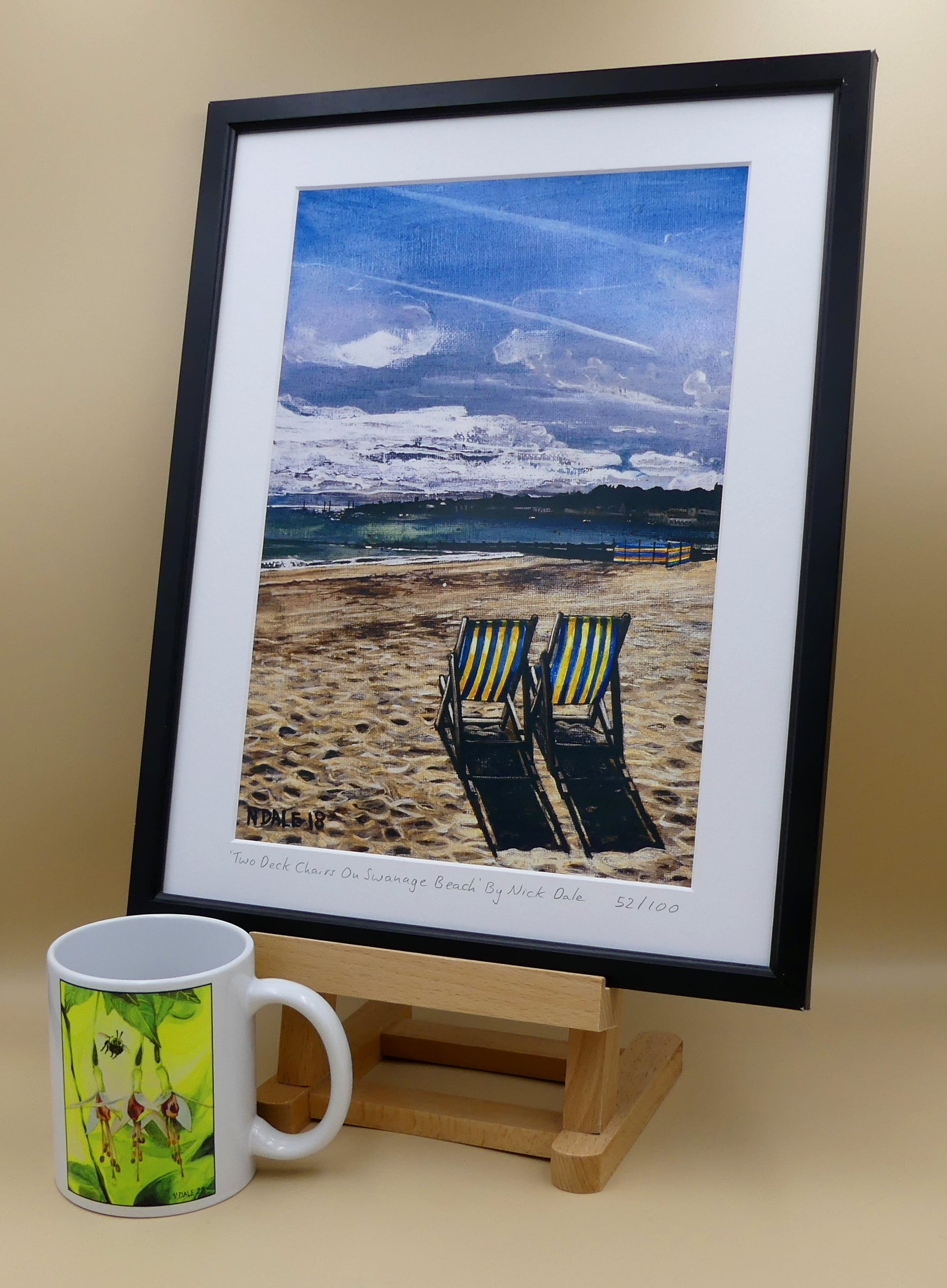 Image of a Medium Art Print featuring a copy of the painting 'Two Deck Chairs On Swanage Beach' by Nick Dale. The print depicts  two deck chairs overlooking the calm water from Swanage Beach (Dorset) towards Peveril Point. The print is situated on a small easel in a black frame next to a mug.