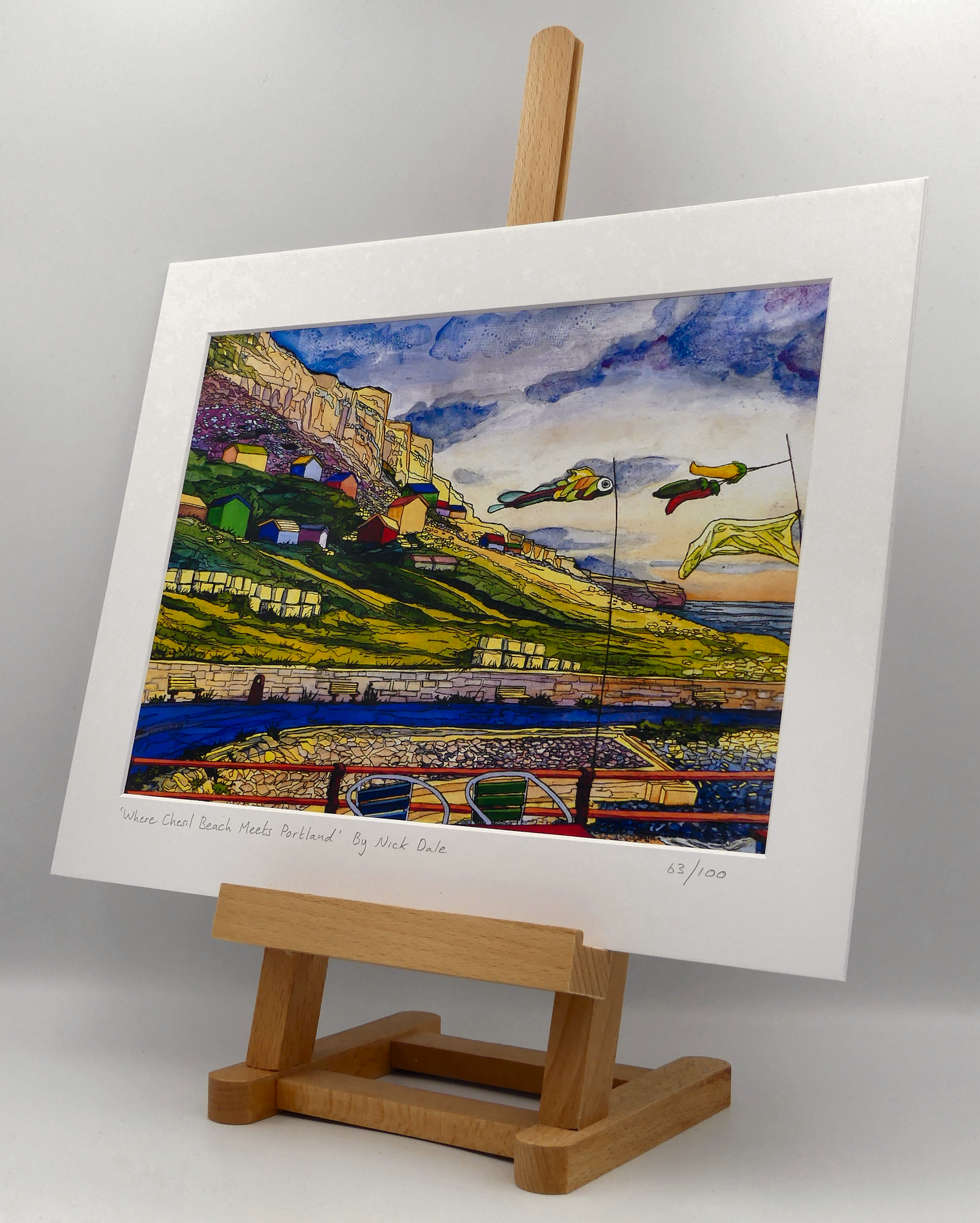 Image of a Medium Art Print featuring a copy of the painting 'Where Chesil Beach Meets Portland' by Nick Dale. The print depicts the point where Chesil Beach meets the Isle of Portland, Dorset. In the foreground are the chairs and decorative flags of Quiddles Café Beach Bar. On the slopes below the cliffs of Portland are the many beach huts which overlook the iconic Chesil Cove. The print is situated on a small easel.
