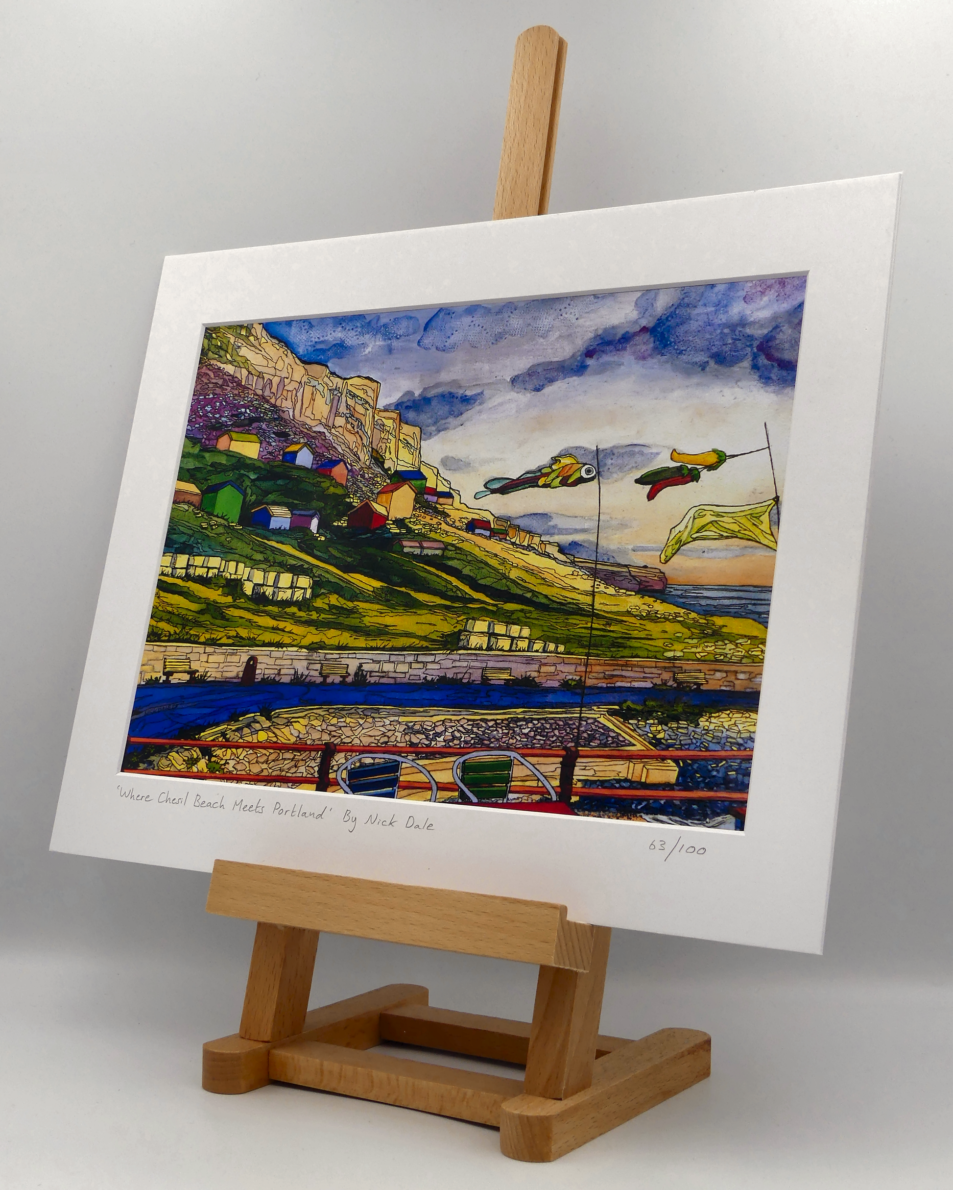 Image of a Medium Art Print featuring a copy of the painting 'Where Chesil Beach Meets Portland' by Nick Dale. The print depicts the point where Chesil Beach meets the Isle of Portland, Dorset. In the foreground are the chairs and decorative flags of Quiddles Café Beach Bar. On the slopes below the cliffs of Portland are the many beach huts which overlook the iconic Chesil Cove. The print is situated on a small easel.