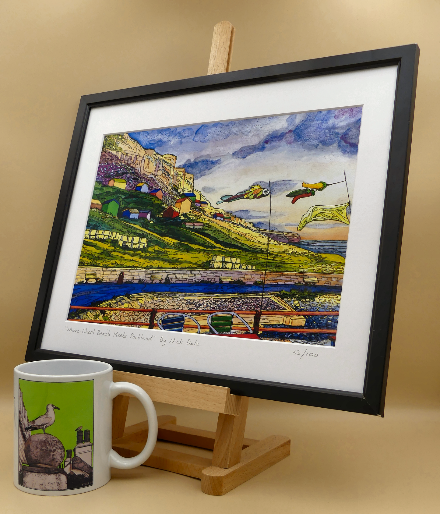 Image of a Medium Art Print featuring a copy of the painting 'Where Chesil Beach Meets Portland' by Nick Dale. The print depicts the point where Chesil Beach meets the Isle of Portland, Dorset. In the foreground are the chairs and decorative flags of Quiddles Café Beach Bar. On the slopes below the cliffs of Portland are the many beach huts which overlook the iconic Chesil Cove. The print is situated on a small easel in a black frame next to a mug.
