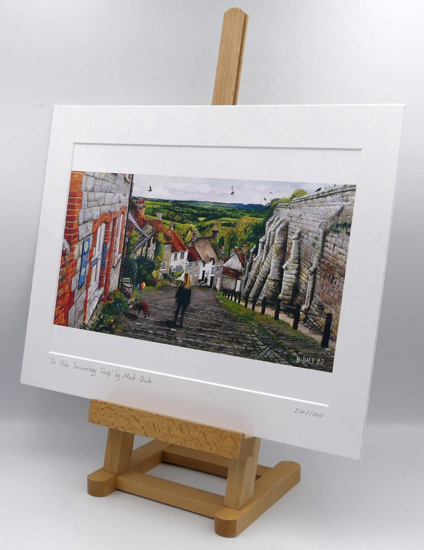 Image of Medium Art Print featuring a copy of the painting 'Ye Olde Technology Shop' by Nick Dale. The print depicts the view down Gold Hill in Shaftesbury, Dorset but in a slightly surreal form as the scene depicts a shop selling helium used to enable dogs to float. The dog is trying to get to a dropped ice-cream cone but is unable to reach as it floats above it. The print is situated on a small easel.