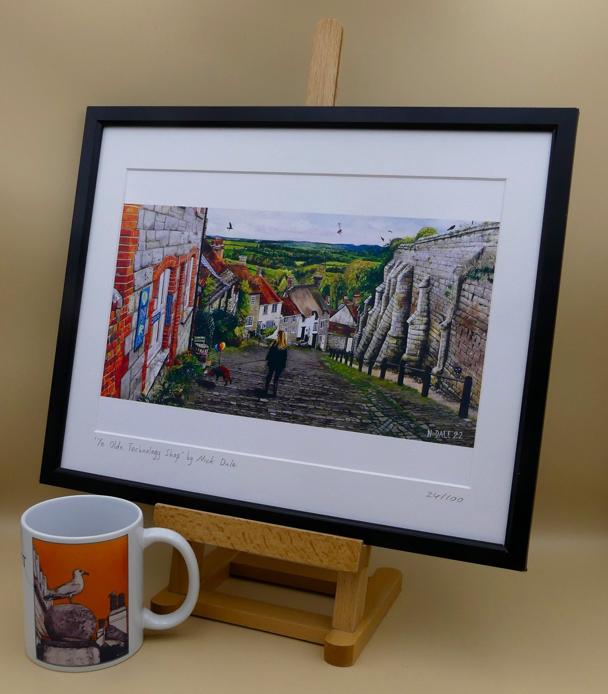 Image of Medium Art Print featuring a copy of the painting 'Ye Olde Technology Shop' by Nick Dale. The print depicts the view down Gold Hill in Shaftesbury, Dorset but in a slightly surreal form as the scene depicts a shop selling helium used to enable dogs to float. The dog is trying to get to a dropped ice-cream cone but is unable to reach as it floats above it. The print is situated in a black frame next to a mug.
