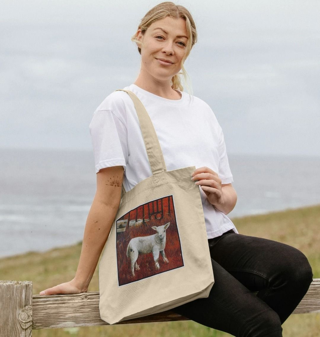 Image of a female model carrying a Natural Tote Bag featuring a modified copy of a detail from the painting 'The Gate' by Nick Dale. The bag depicts a lamb in front of an ornamental garden gate reused by a farmer somewhere in North Devon, probably to the south of Valley of Rocks, Lynton. The lamb was observed on Lundy Island, Devon.
