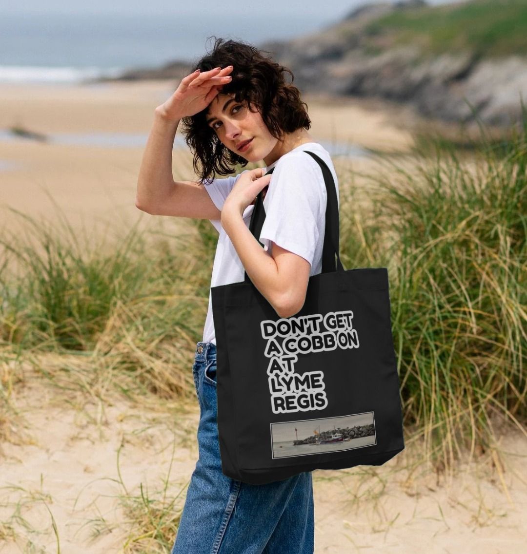 Image of a female model carrying a Black Shopper Tote Bag featuring a copy of the painting 'End Of The Line' by Nick Dale with the slogan 'Don't Get A Cobb On At Lyme Regis' written above it. The bag depicts a fisherman preparing his catch for unloading at Lyme Regis as the birds line up on the breakwater and patiently await their turn to feed.