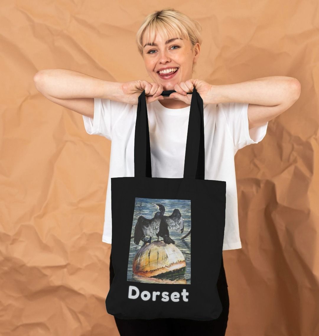 Image of a female model holding a Black Tote Bag featuring a modified copy of the painting 'Cormorant At West Bay' by Nick Dale. The bag depicts a close-up of a cormorant sunbathing on a mooring buoy in West Bay's harbour in Dorset.