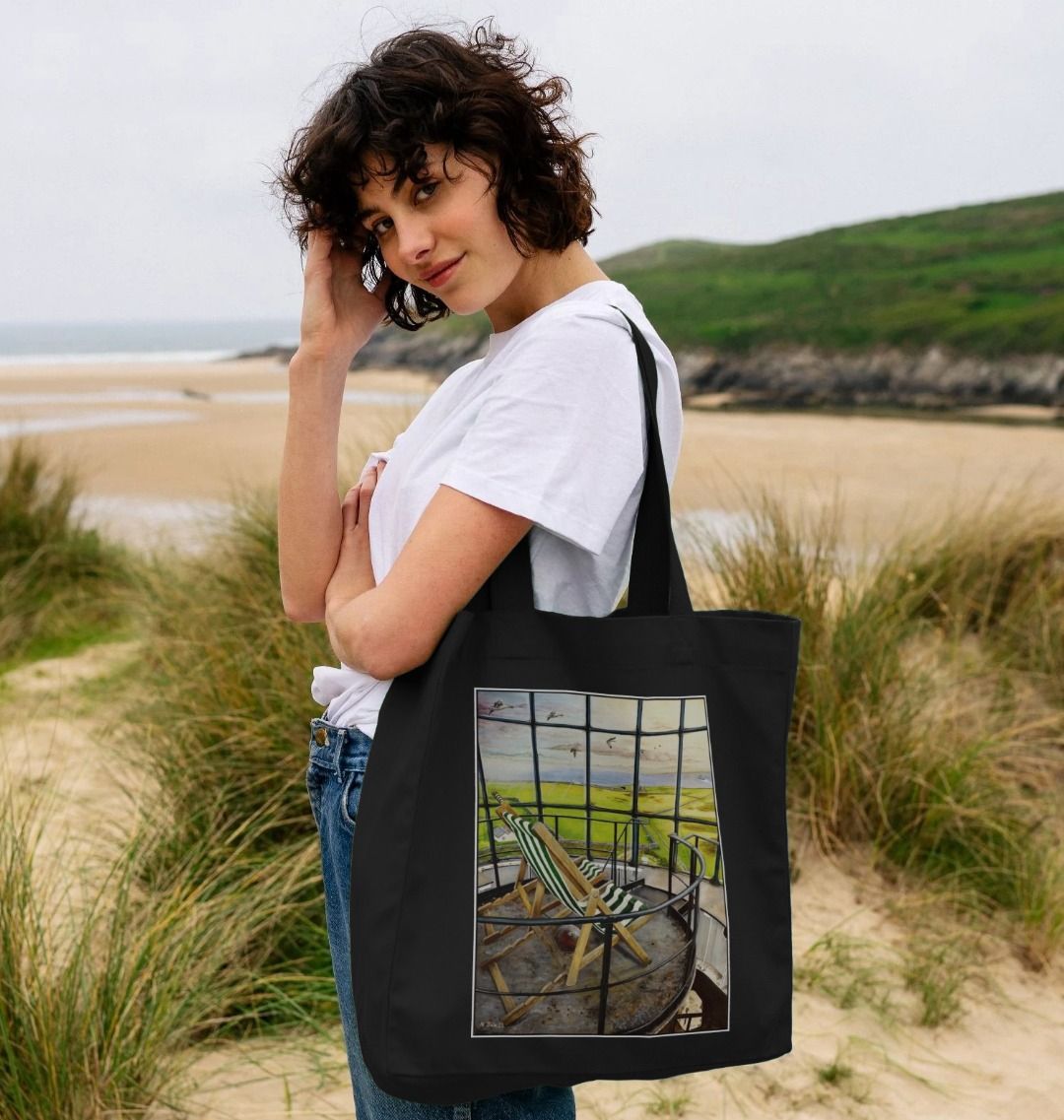 Image of a female model carrying a Black Shopper Tote Bag featuring a copy of the painting 'Let's Be Together Alone' by Nick Dale. The bag depicts the view from the lantern room of the Old Light lighthouse on Beacon Hill, on the island of Lundy, Devon.