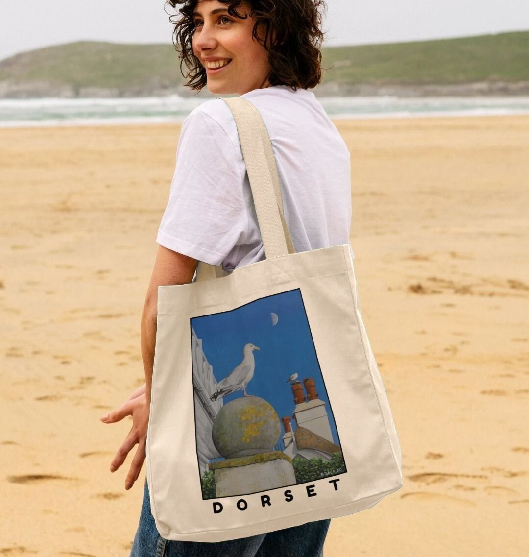Image of female model holding Natural Shopper Tote Bag featuring a copy of the painting 'A View Of The Sea At Lyme Regis' by Nick Dale. The bag depicts a gull and buildings along Marine Parade, Lyme Regis.