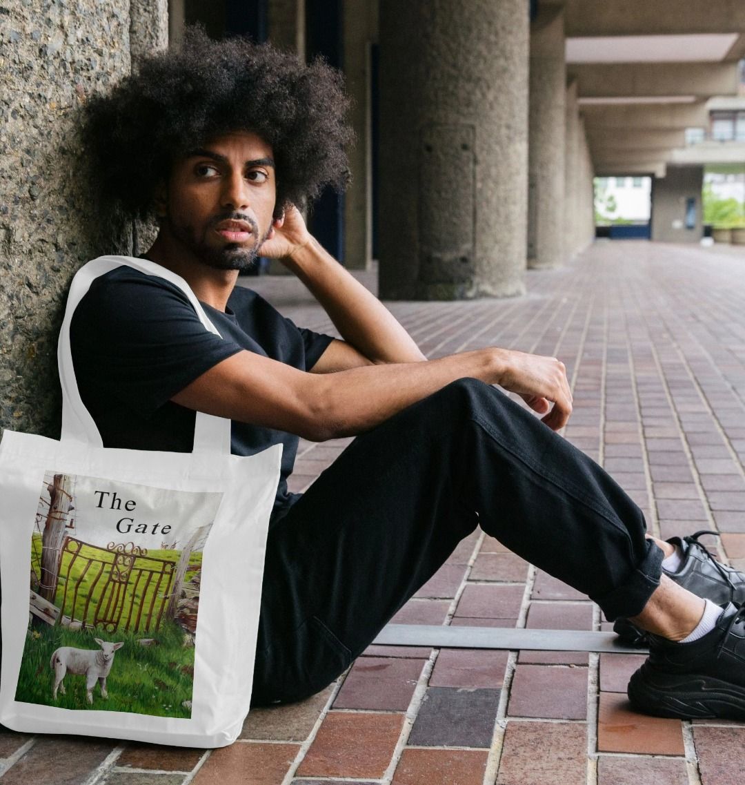 Image of a male model carrying a White Tote Bag featuring a copy of a detail from the painting 'The Gate' by Nick Dale, with the words 'the Gate' written above the gate. The bag depicts a lamb in front of an ornamental garden gate reused by a farmer somewhere in North Devon, probably to the south of Valley of Rocks, Lynton. The lamb was observed on Lundy Island, Devon.