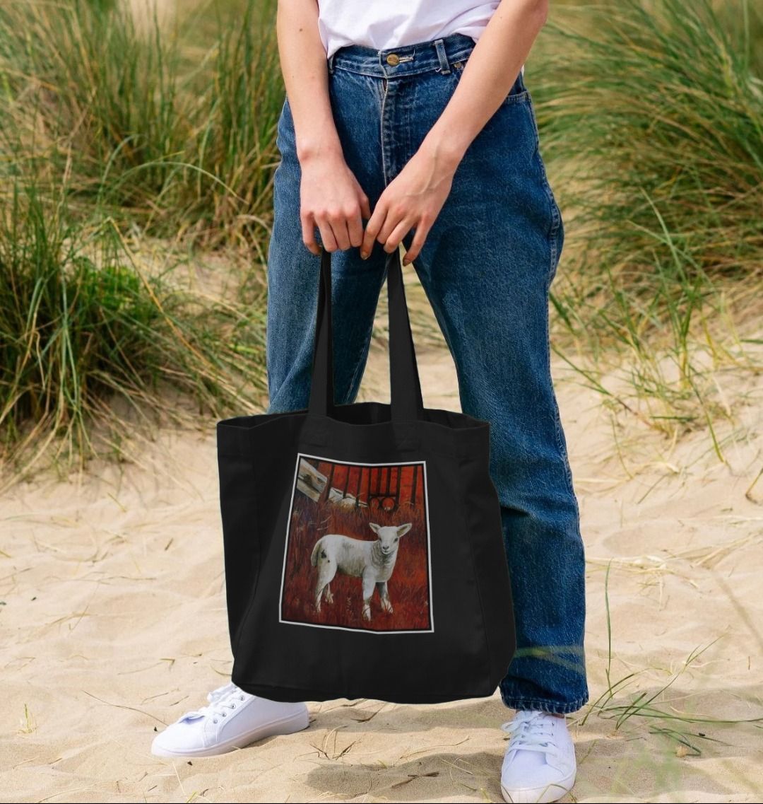 Image of a female model holding a Black Shopper Tote Bag featuring a modified copy of a detail from the painting 'The Gate' by Nick Dale. The bag depicts a lamb in front of an ornamental garden gate reused by a farmer somewhere in North Devon, probably to the south of Valley of Rocks, Lynton. The lamb was observed on Lundy Island, Devon.