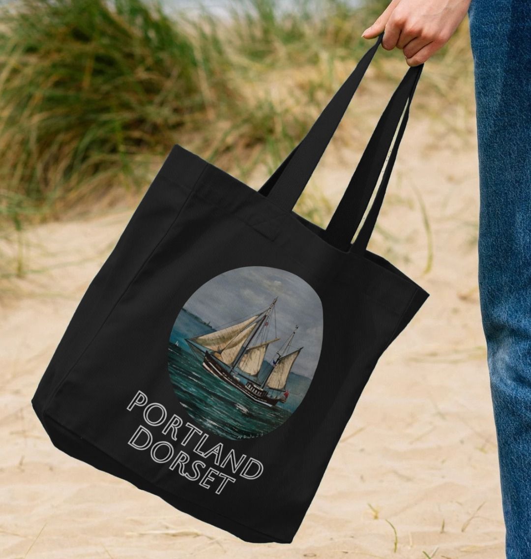 Image of a female model carrying a Black Shopper Tote Bag featuring a copy of the painting 'Moonfleet Sailing Past Portland' by Nick Dale, with the words 'Portland Dorset' written beneath it. The bag depicts the tall ship Moonfleet sailing past the western coast of Portland, Dorset. As observed from Ringstead Beach.