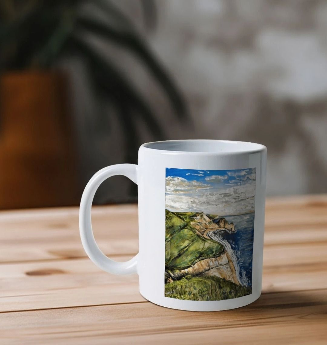 Image of a White Mug on a table featuring a copy of the painting 'Durdle Door From The West' by Nick Dale. The handle is orientated to the left. The mug depicts a view along the Jurassic Coast to the famous Dorset limestone arch landmark, as seen from the coastal path to the west.