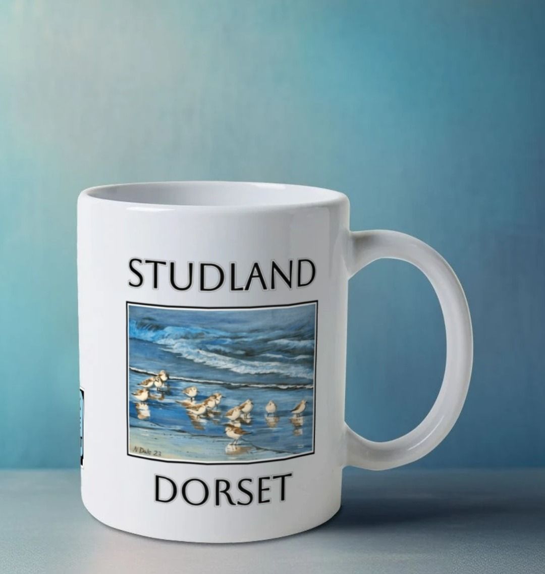 Image of a White Mug on a white table featuring a copy of the painting 'Studland Misty Morning' by Nick Dale, with the word 'Studland' written above the picture and 'Dorset' written beneath it. The handle is orientated to the right. The mug depicts Dunlins on Studland Beach (Dorset) on a crisp and misty morning.