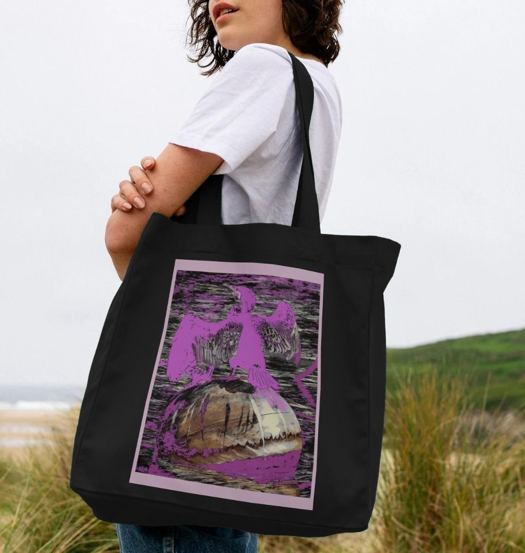 Image of a female model holding a Black Shopper Tote Bag featuring a modified copy of the painting 'Cormorant At West Bay' by Nick Dale. The bag depicts a close-up of a cormorant sunbathing on a mooring buoy in West Bay's harbour in Dorset.