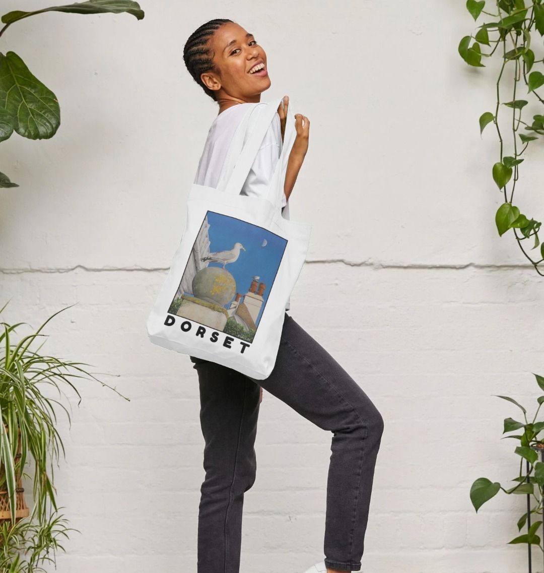 Image of female model holding White Tote Bag featuring a copy of the painting 'A View Of The Sea At Lyme Regis' by Nick Dale. The bag depicts a gull and buildings along Marine Parade, Lyme Regis.