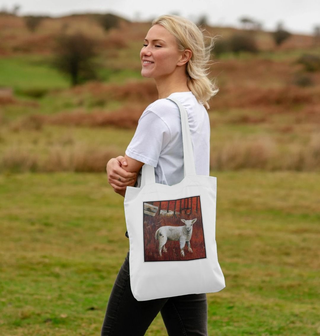 Image of a female model carrying a White Tote Bag featuring a modified copy of a detail from the painting 'The Gate' by Nick Dale. The bag depicts a lamb in front of an ornamental garden gate reused by a farmer somewhere in North Devon, probably to the south of Valley of Rocks, Lynton. The lamb was observed on Lundy Island, Devon.