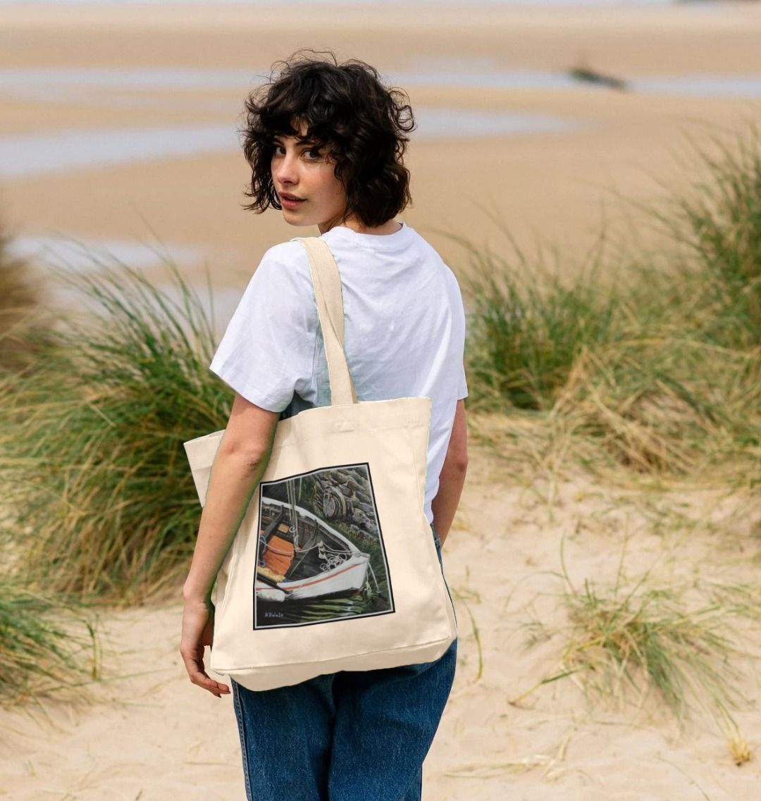 Image of a female model carrying a Natural Shopper Tote Bag featuring a copy of the painting 'The Outlet' by Nick Dale. The bag depicts a view down into the harbour at Lynmouth in North Devon showing a small sailing boat at rest against the harbour wall next to a water outfall pipe.