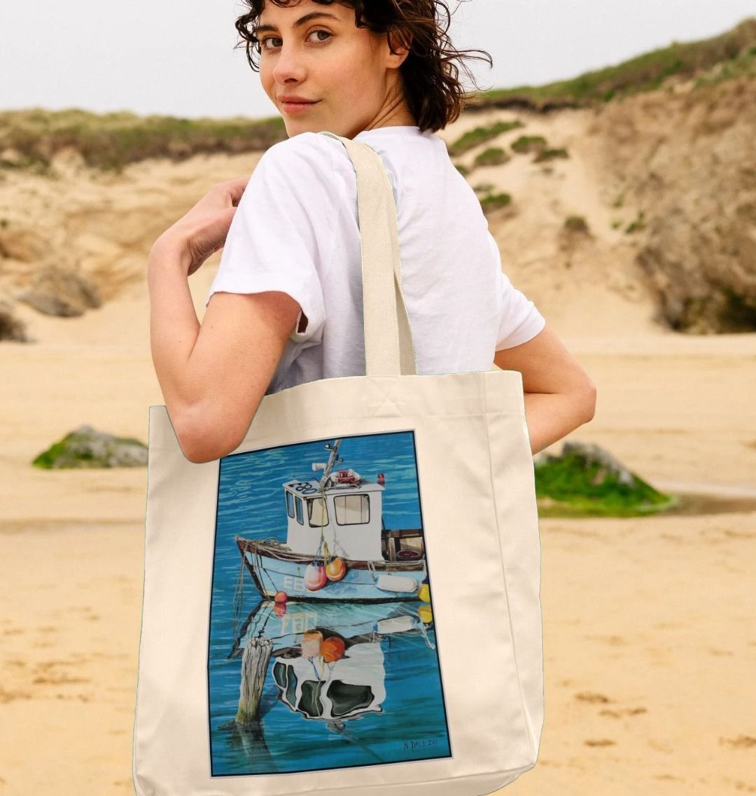Image of a female model carrying a Natural Shopper Tote Bag featuring a copy of the painting 'God's Own 99 Flake Pierces The Veil' by Nick Dale. The bag depicts a fishing boat at rest within the harbour of Lyme Regis, Dorset. The title is based on the similarity of the wooden pile to a chocolate flake as inserted into an ice cream cone. This seemed appropriate given the draw of the town to tourists.