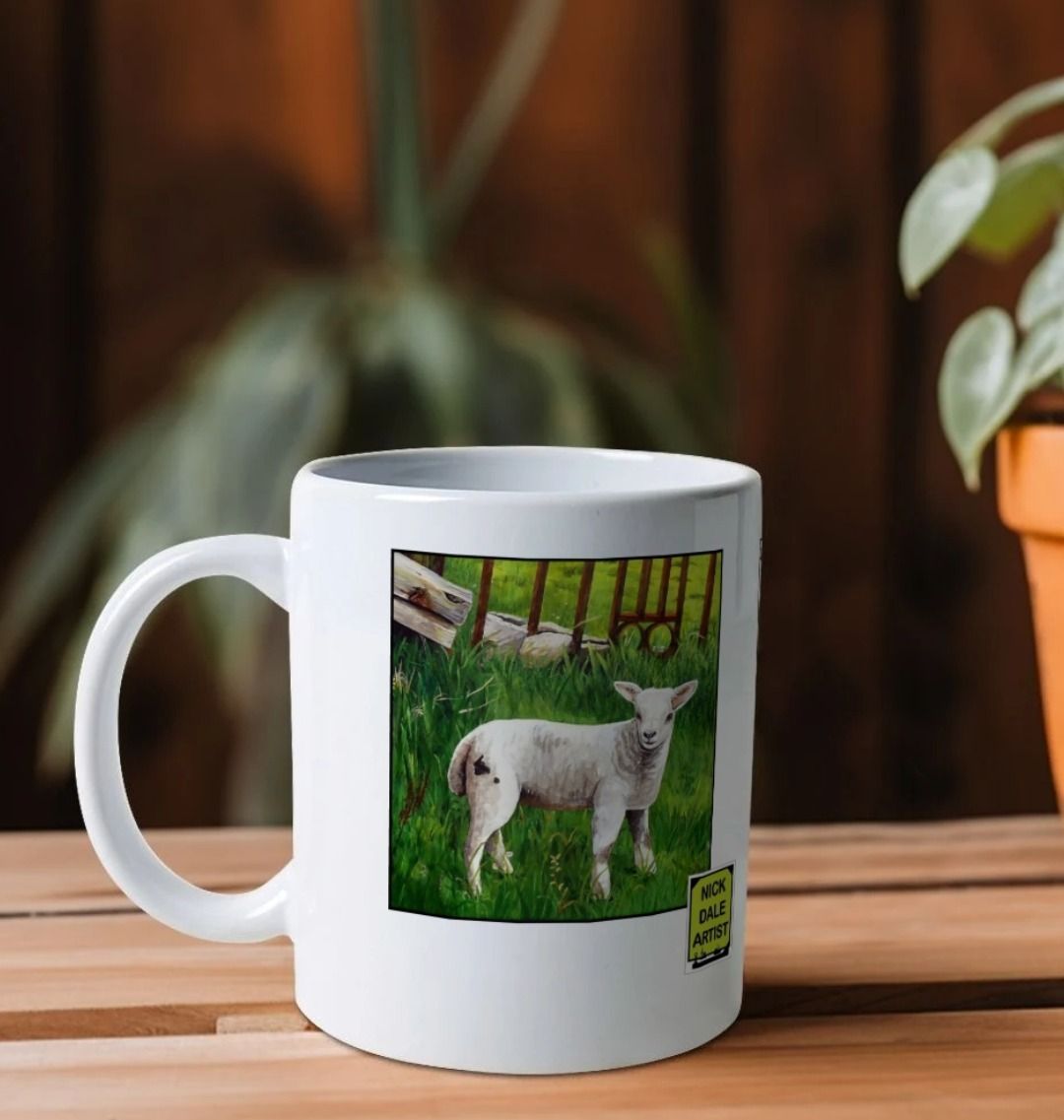 Image of a White Mug on a wooden shelf featuring a copy of the painting 'The Gate' by Nick Dale; as well as a close-up detail of a lamb from the same painting. Image also shows the artist's logo on the mug. The handle is orientated to the left. The mug depicts an ornamental garden gate reused by a farmer somewhere in North Devon, probably to the south of Valley of Rocks, Lynton. The animals depicted are all based on animals observed on Lundy Island, Devon.