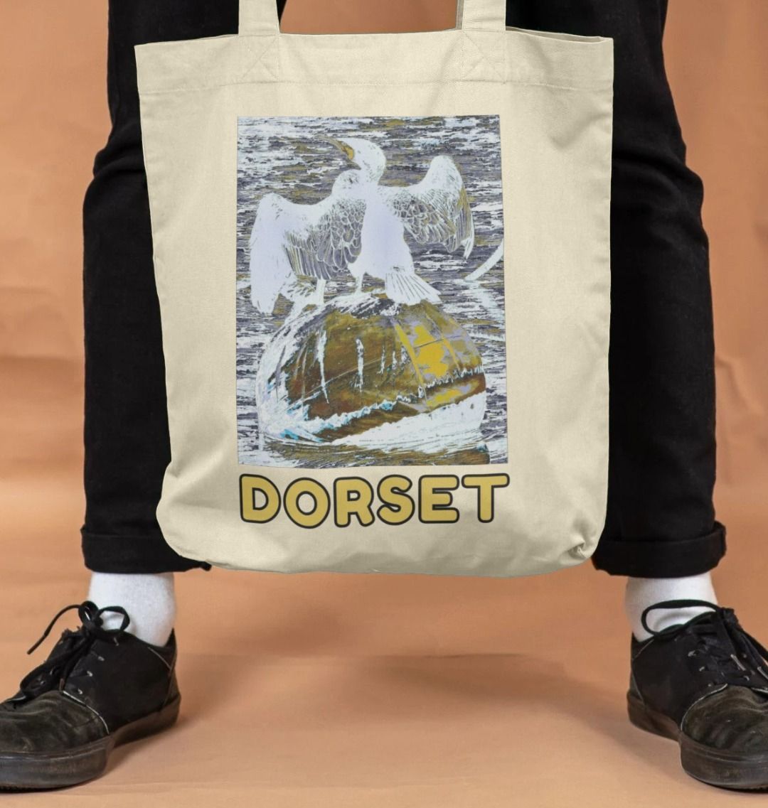 Image of a male model holding a Natural Tote Bag featuring a modified copy of the painting 'Cormorant At West Bay' by Nick Dale. The bag depicts a close up of a cormorant sunbathing on a mooring buoy in West Bay's harbour in Dorset.