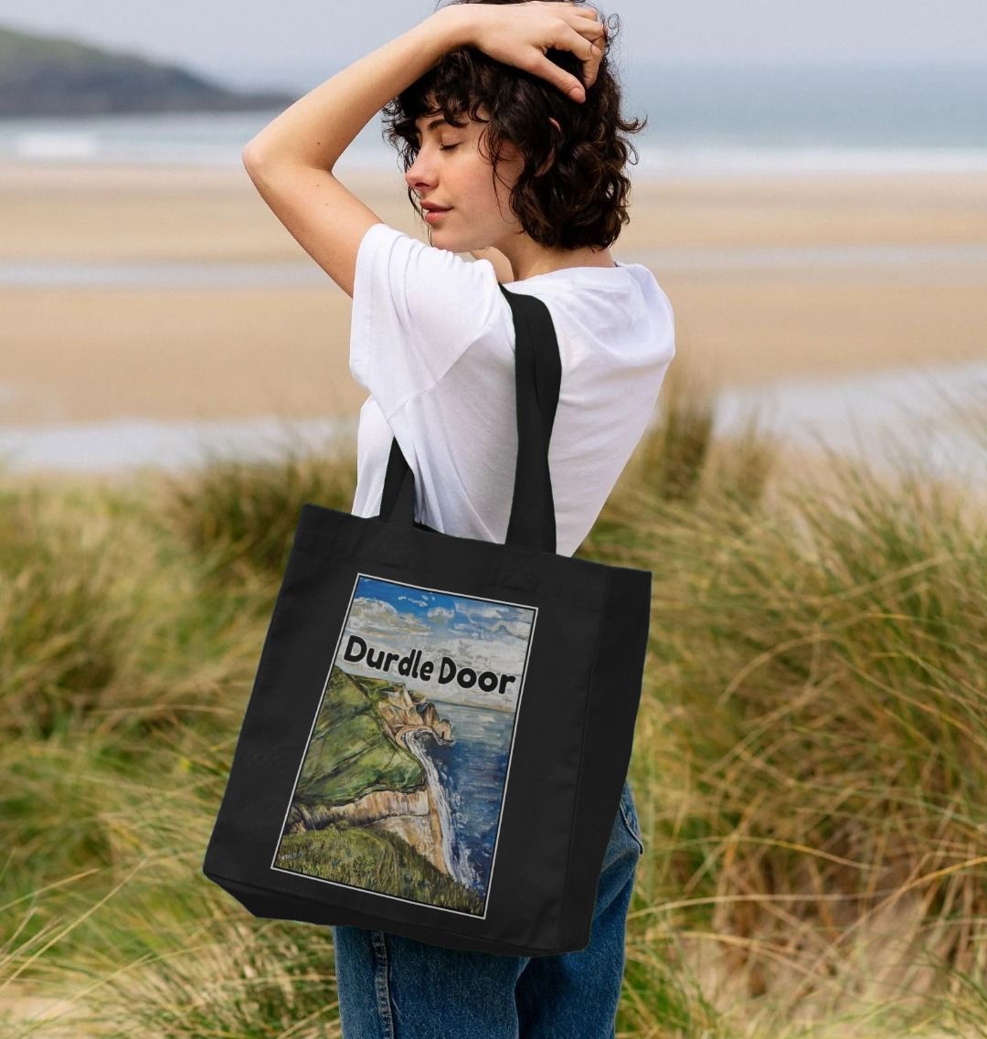 Image of female model carrying a Black Shopper Tote Bag featuring a copy of the painting 'Durdle Door From The West' by Nick Dale with the words 'Durdle Door' written across it. The bag depicts a view along the Jurassic Coast to the famous Dorset limestone arch landmark, as seen from the coastal path to the west.