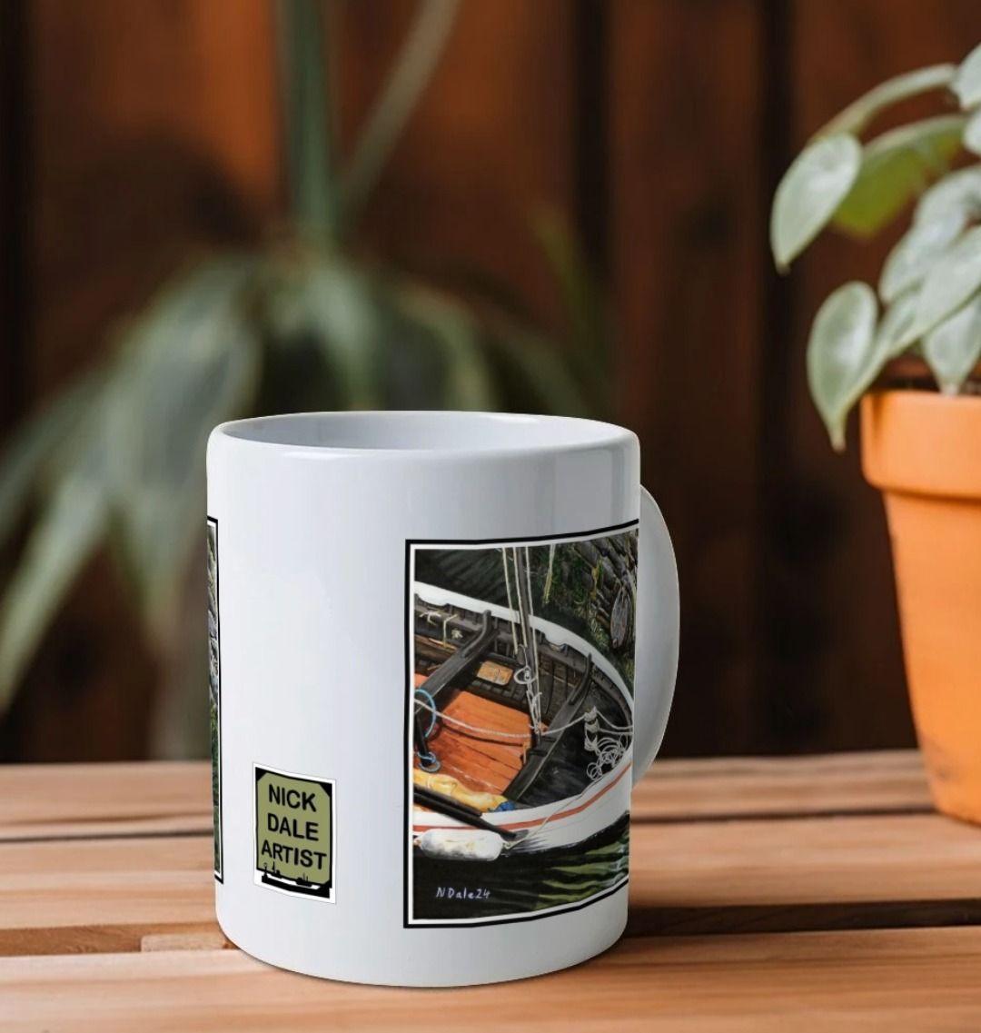 Image of a White Mug featuring a copy of the painting 'The Outlet' by Nick Dale. Image also shows artist's logo on the mug. The mug handle is obscured behind the mug. The mug depicts a view down into the harbour at Lynmouth in North Devon showing a small sailing boat at rest against the harbour wall next to a water outfall pipe.