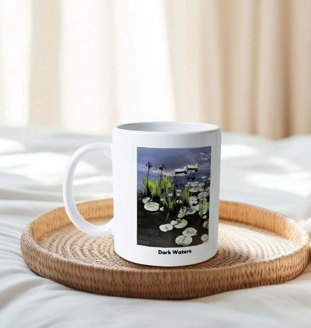 Image of a White Mug on a tray on a bed featuring a copy of the painting 'Dark Waters' by Nick Dale, with the words 'Dark Waters' written beneath it. The handle is orientated to the left. The mug depicts lily pads, irises and ducks on a pond at Knightshayes Court, Devon.