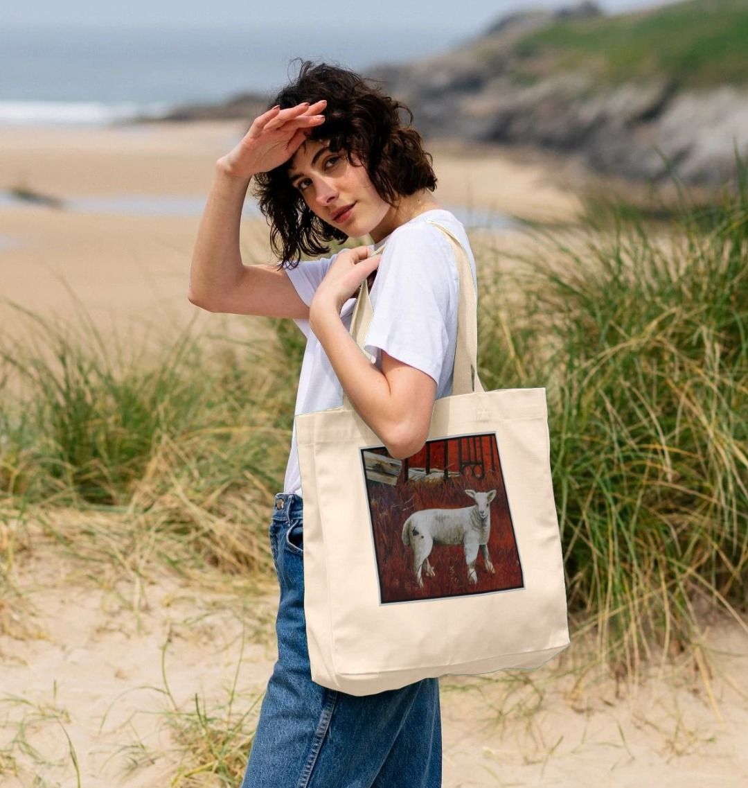 Image of a female model carrying a Natural Shopper Tote Bag featuring a modified copy of a detail from the painting 'The Gate' by Nick Dale. The bag depicts a lamb in front of an ornamental garden gate reused by a farmer somewhere in North Devon, probably to the south of Valley of Rocks, Lynton. The lamb was observed on Lundy Island, Devon.